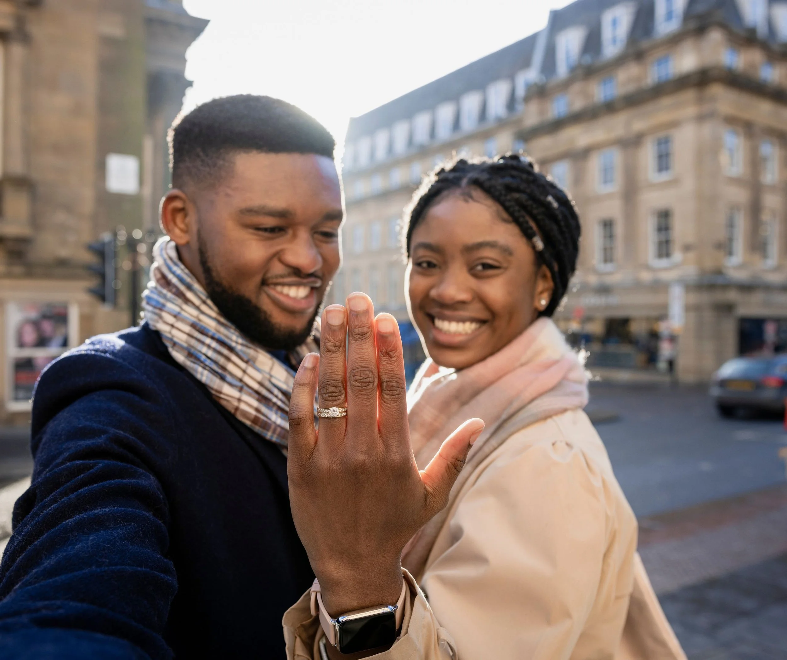 Un couple souriant montrant une bague de fiançailles en pleine rue avec bâtiments anciens en arrière-plan.