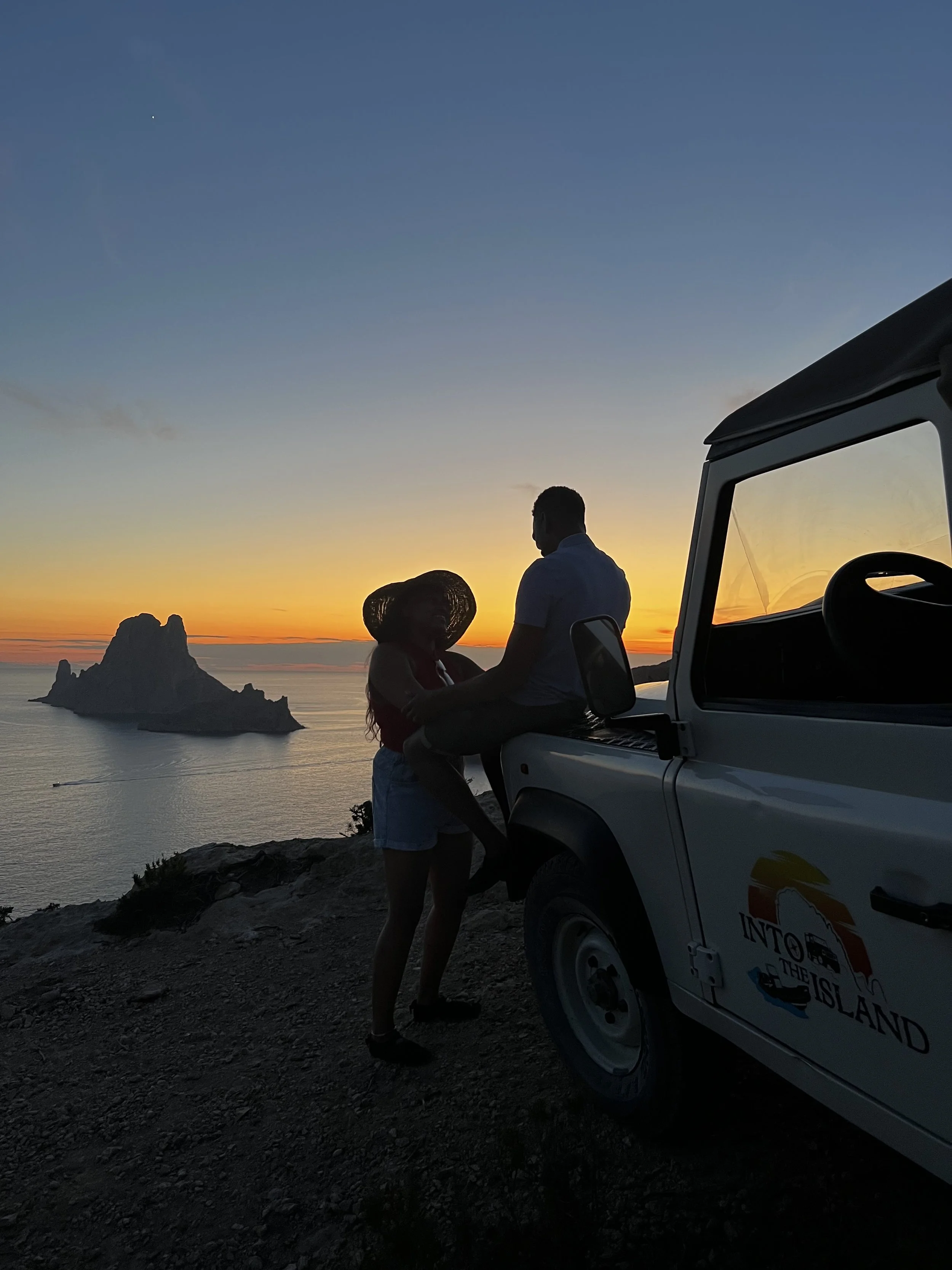 Deux personnes, une femme portant un chapeau, et un homme, debout près d'un véhicule avec une vue sur la mer, lors d'un coucher de soleil.