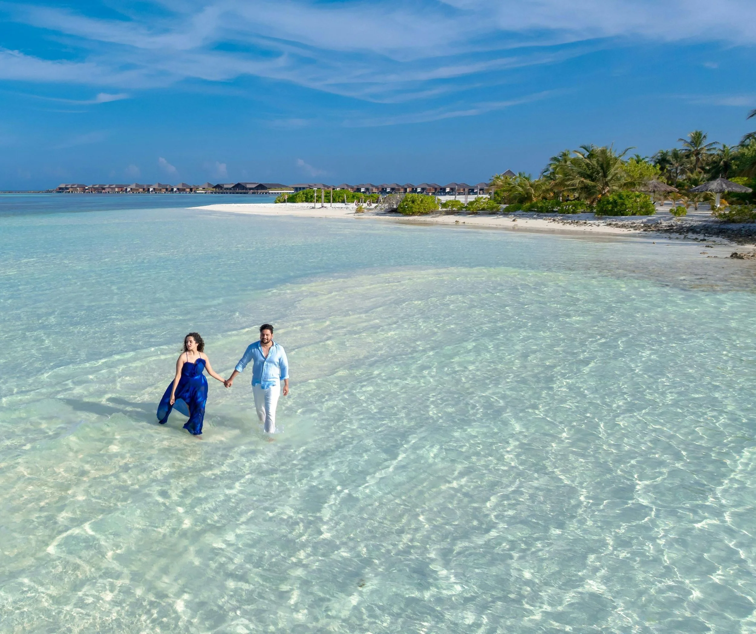 Un couple marche main dans la main dans une mer cristalline, avec des villas sur pilotis au loin et un ciel bleu clair.
