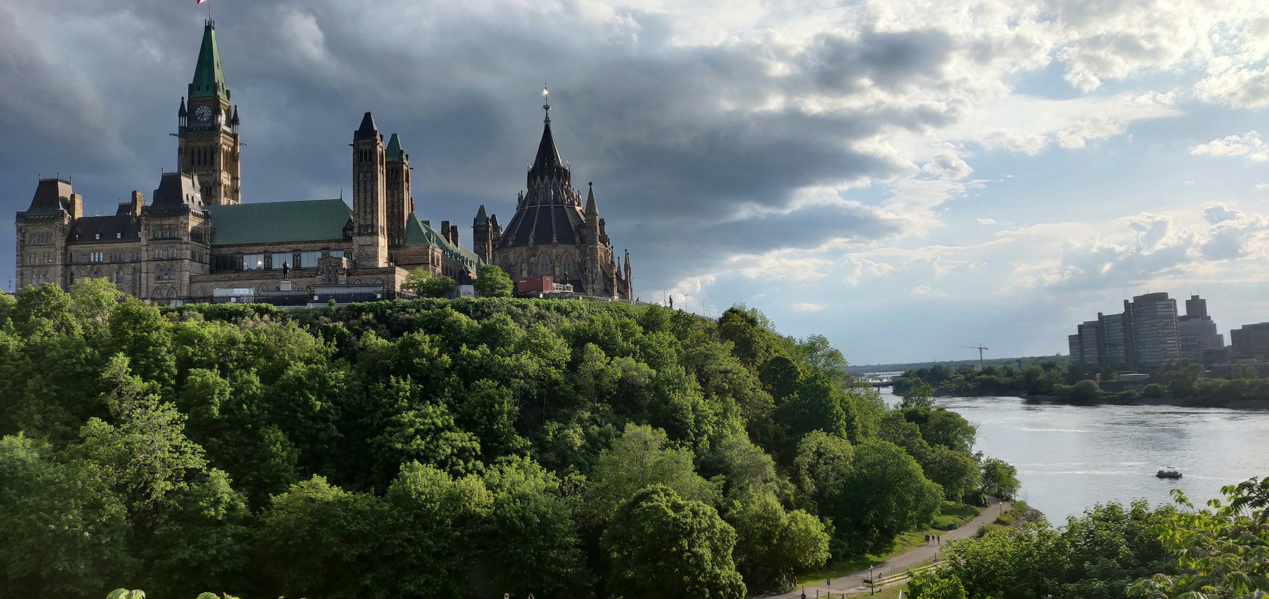 View of a large historic castle-like building with tall spires and towers, situated on a hill covered with lush green trees, next to a river with a boat, under a cloudy sky.