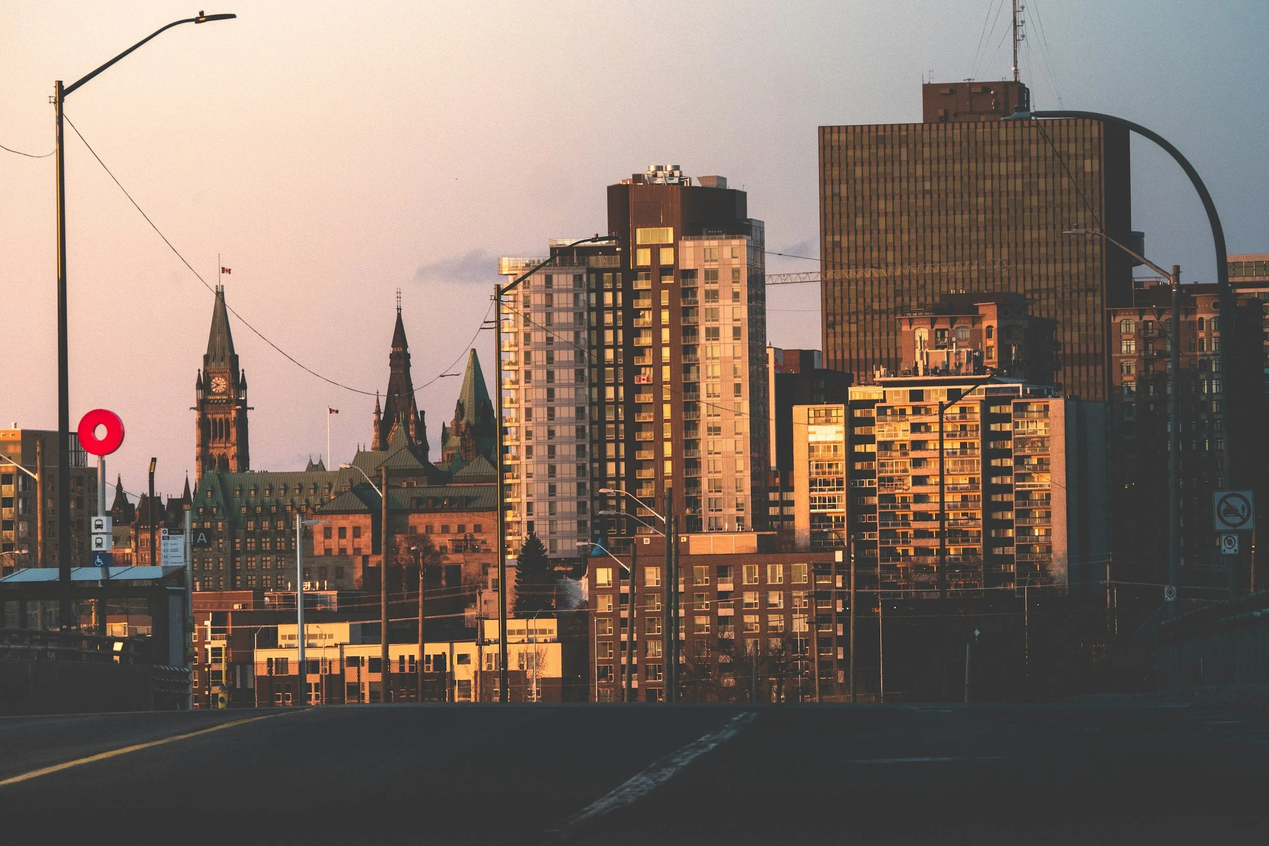City skyline at sunset with a mix of modern high-rise buildings and historic church steeples, viewed from a roadway with streetlights.