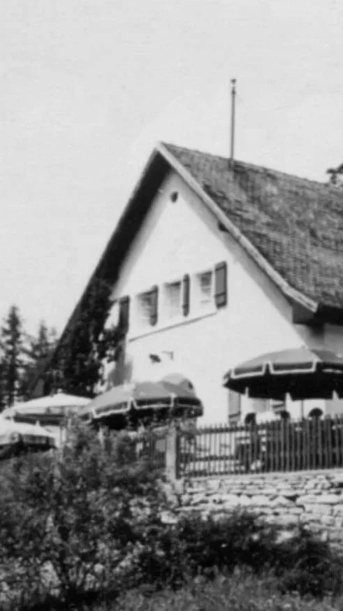 Ein weißes Haus mit Balkon, Dach und mehreren Sonnenschirmen im Außenbereich, umgeben von einer kleinen Gartenmauer und Büschen, im Hintergrund Bäume.