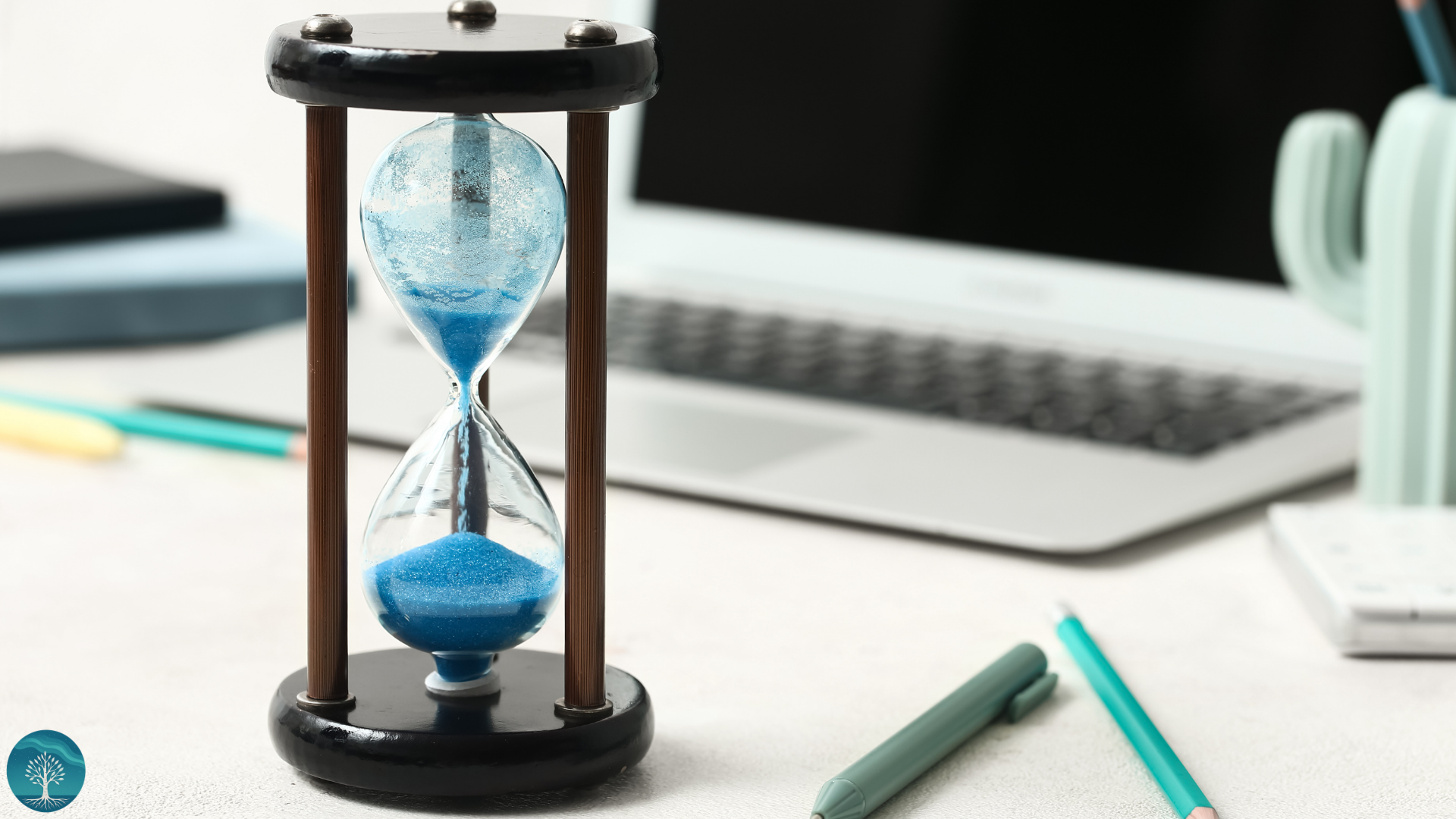 An hourglass with blue sand on a white desk, surrounded by markers, a laptop, and office supplies.