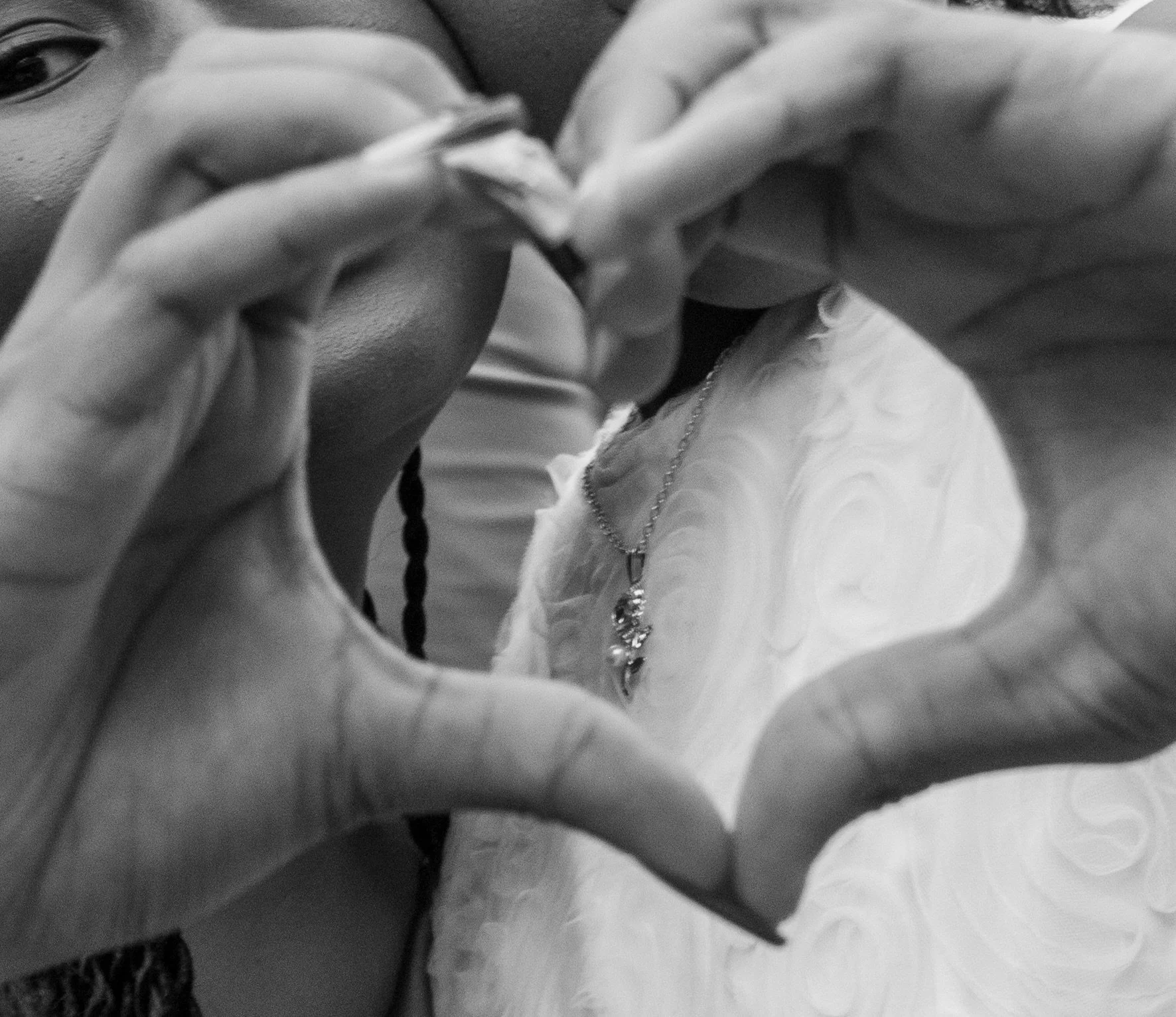A close-up black and white photo of a woman making a heart shape with her hands around her face, focusing on her eye and hand gestures.