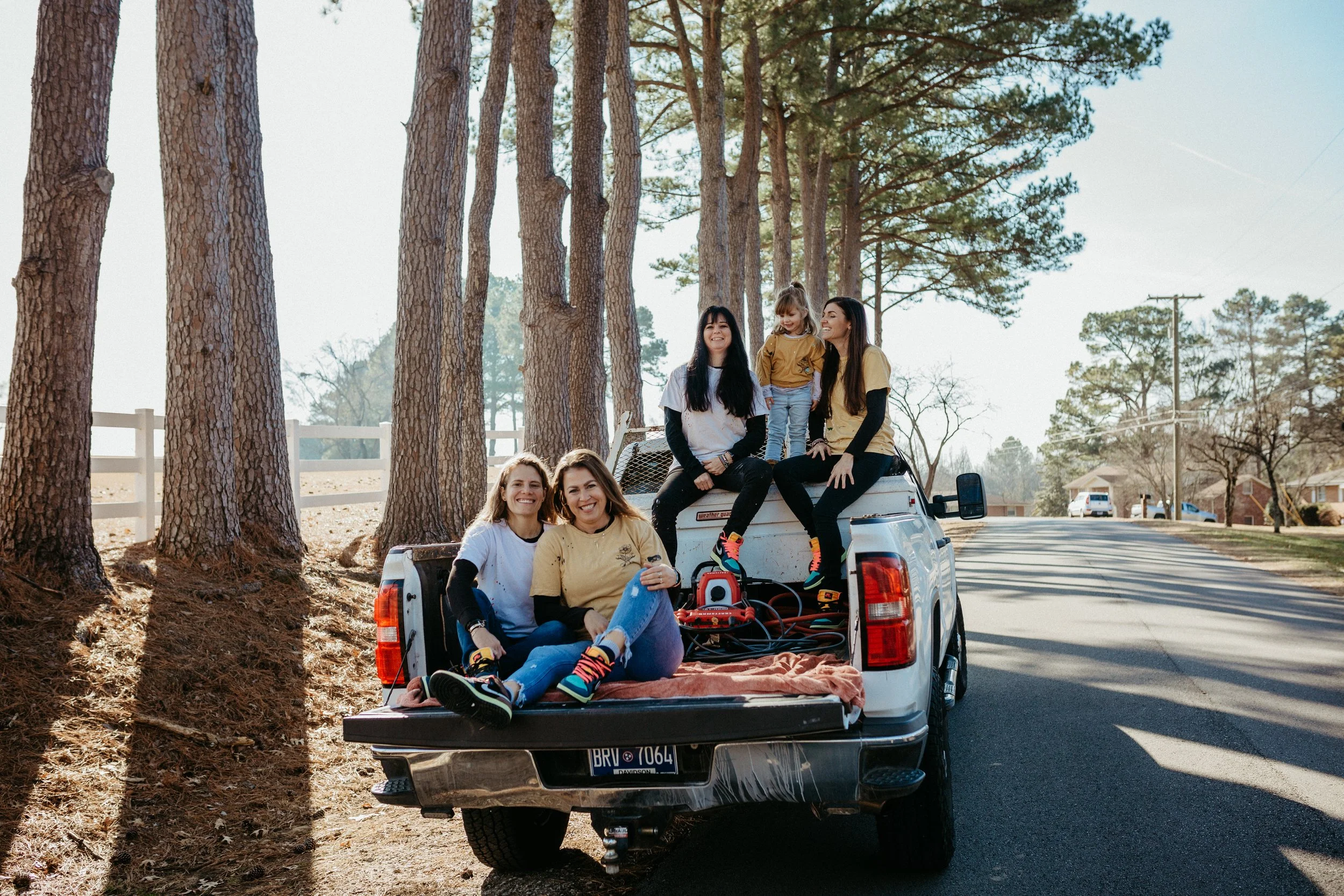 Five women and a young girl are sitting and standing on the back of a pickup truck parked on a residential street surrounded by trees.