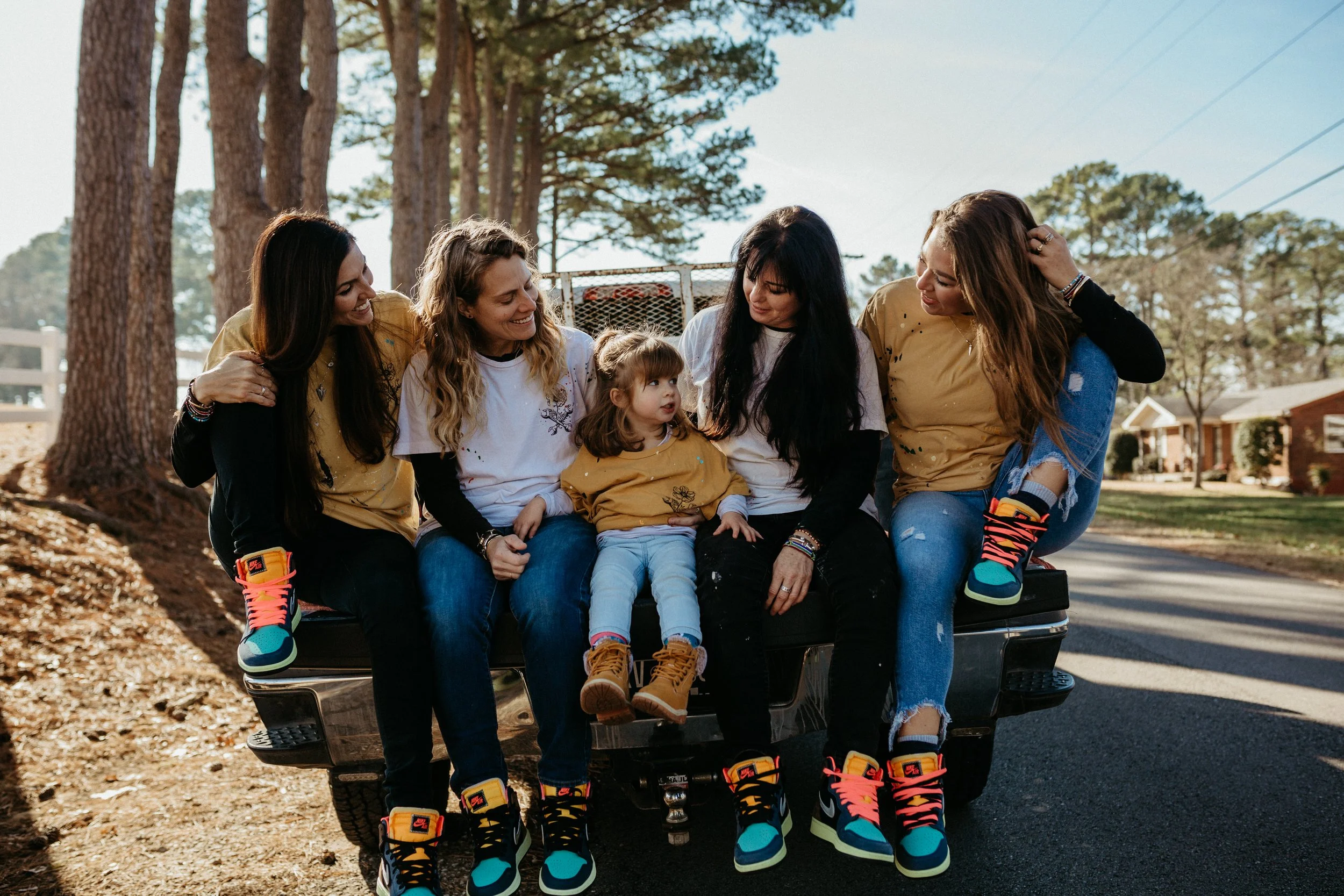 Five women and a young girl sitting together on a vehicle, wearing matching yellow shirts and colorful sneakers, outdoors in a rural setting with trees and houses in the background.