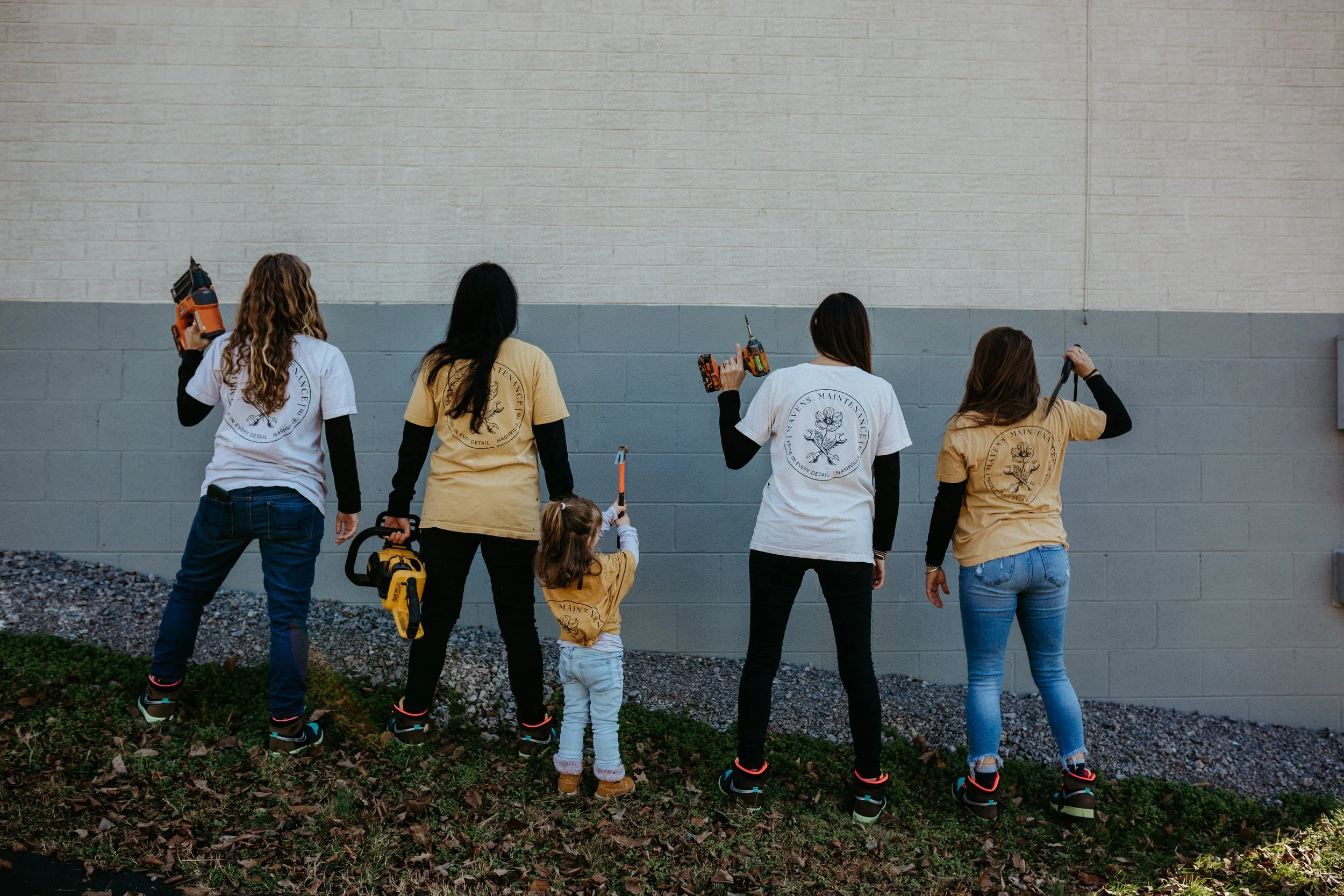 Five women and one young girl working on a wall with power tools, wearing work-related shirts.