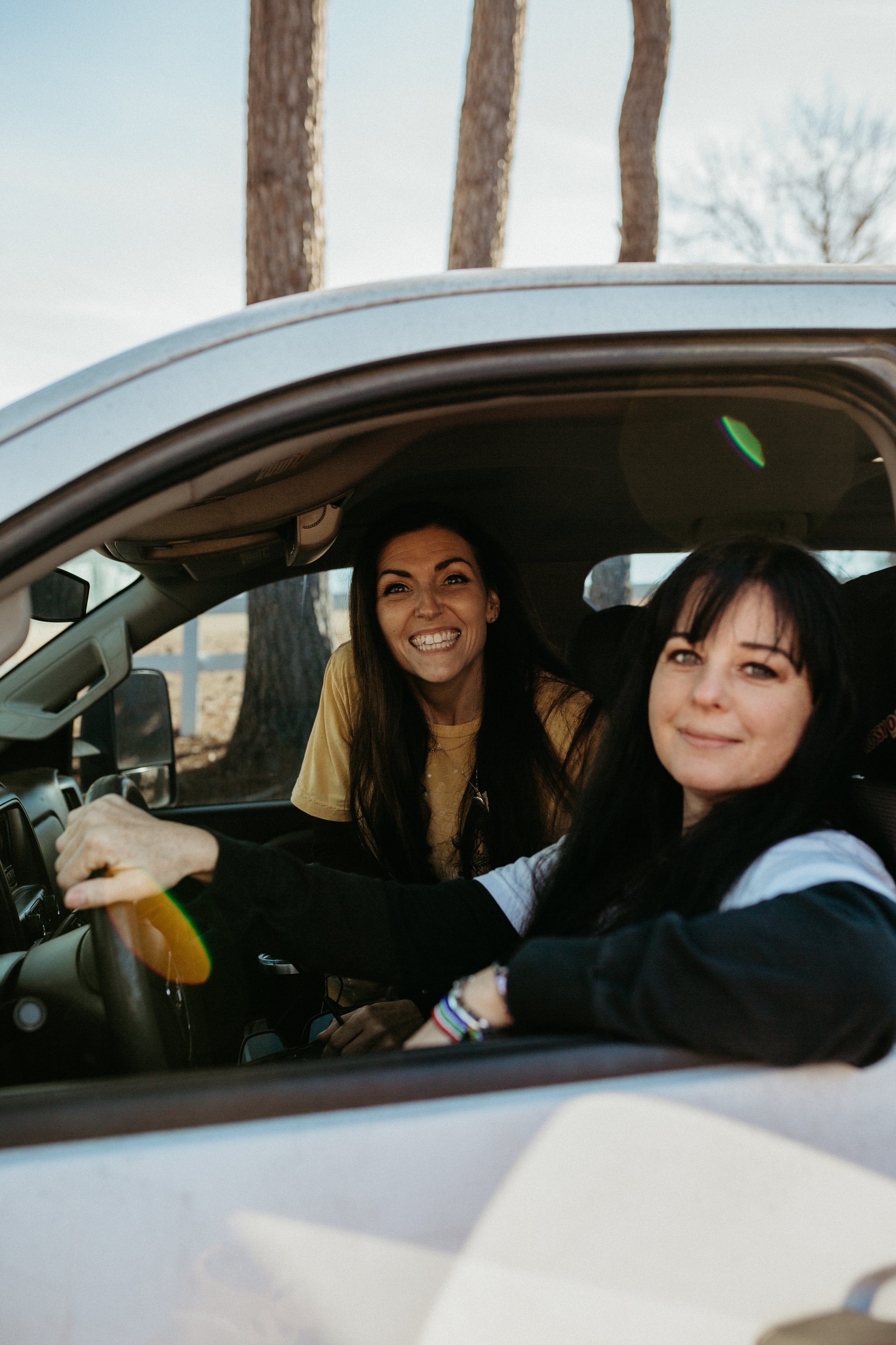Two women inside a white vehicle, smiling at the camera. One woman with long dark hair is in the passenger seat, and another woman with long dark hair in a yellow shirt is leaning inside from the driver's side, outdoors with trees in the background.