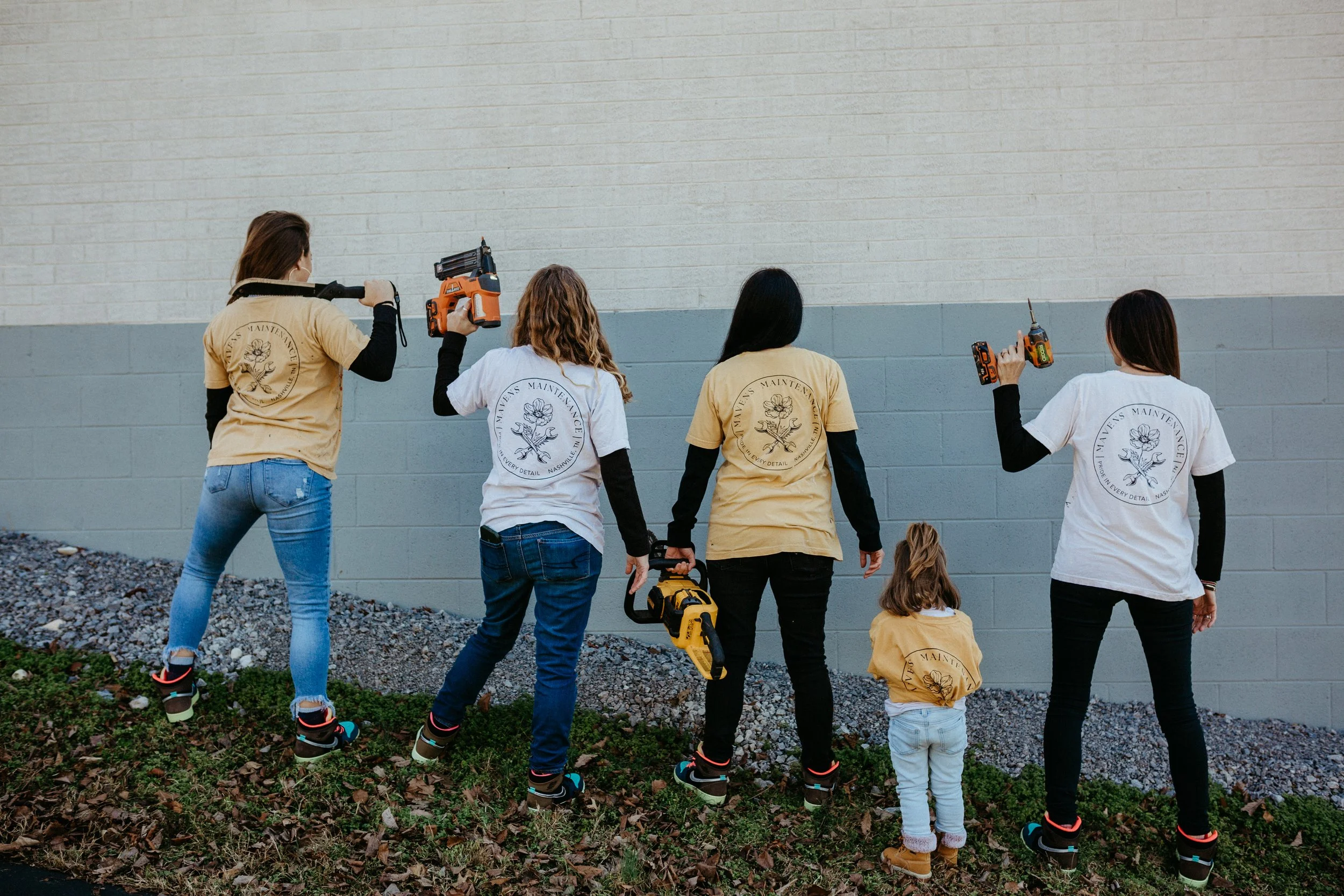 Five women and one young girl working on a home improvement project, wearing matching T-shirts with a flower design on the back, standing outside on a grassy area near a gray brick wall, holding power tools, with some of them wearing safety gear.