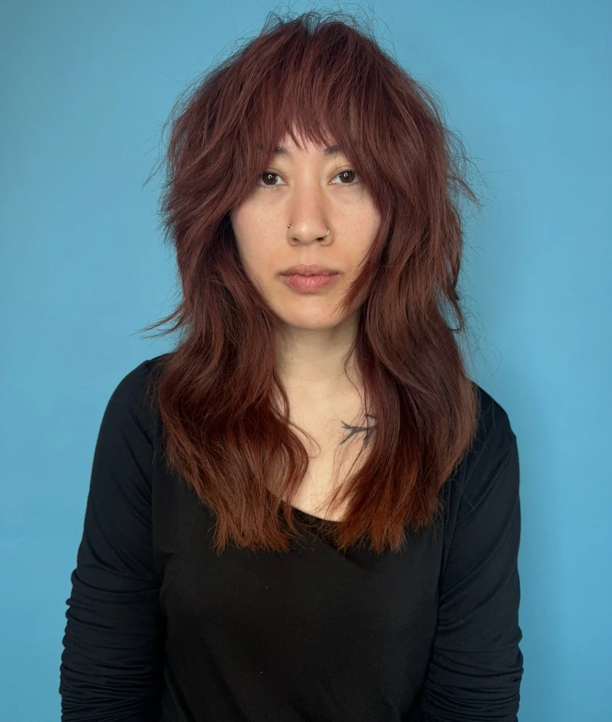 A woman with reddish-brown, wavy hair, wearing a black top, standing against a blue background.