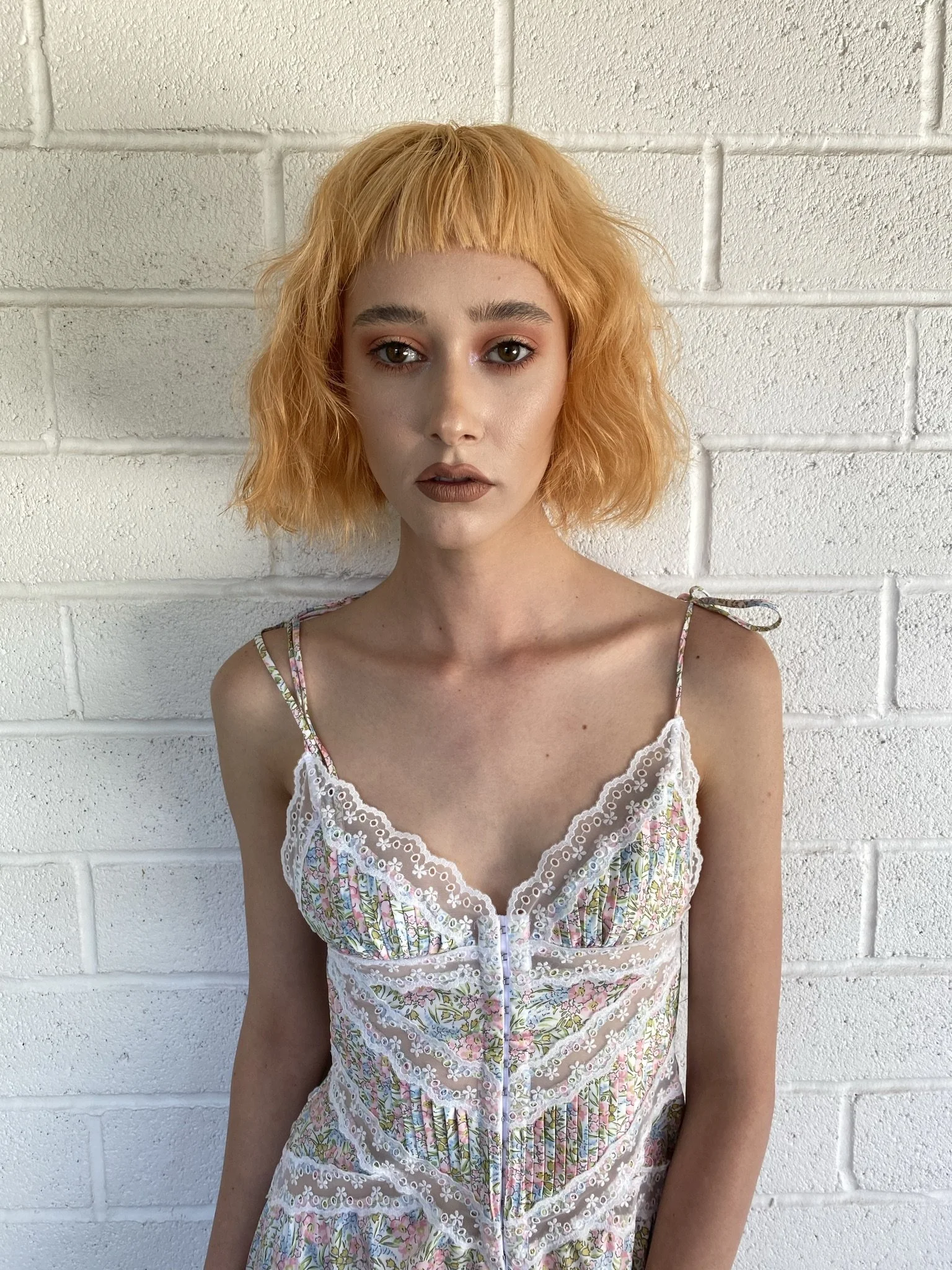 A young woman with short, wavy, ginger hair and makeup standing against a white brick wall.