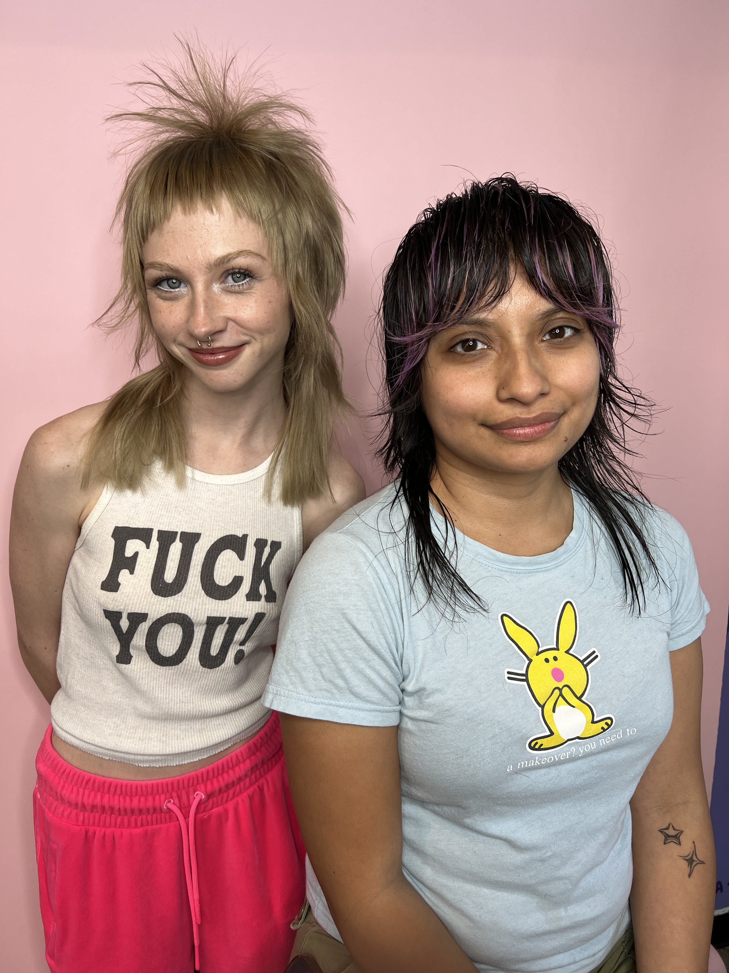 Two female-presenting people with new haircuts. One with long blonde hair and one is a person of color with long dark hair. The blonde is wearing a shirt that says, "Fuck You!"