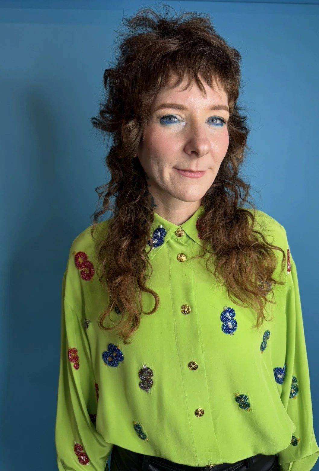 A woman with wavy brown hair and blue eyes wearing a bright lime green blouse decorated with colorful dollar signs and gold buttons, standing against a blue background.