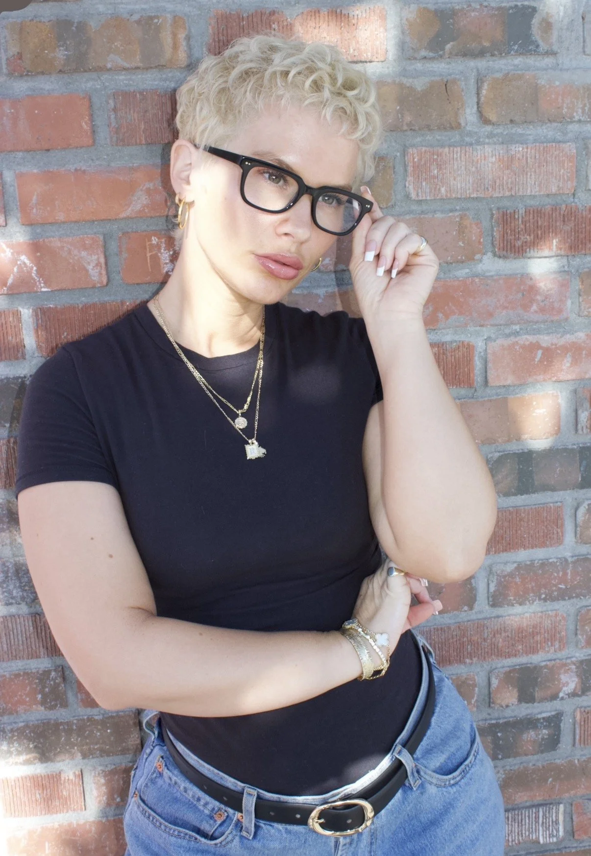 A woman with short blonde curly hair, wearing black glasses, gold jewelry, a black t-shirt, and blue jeans, posing against a brick wall.