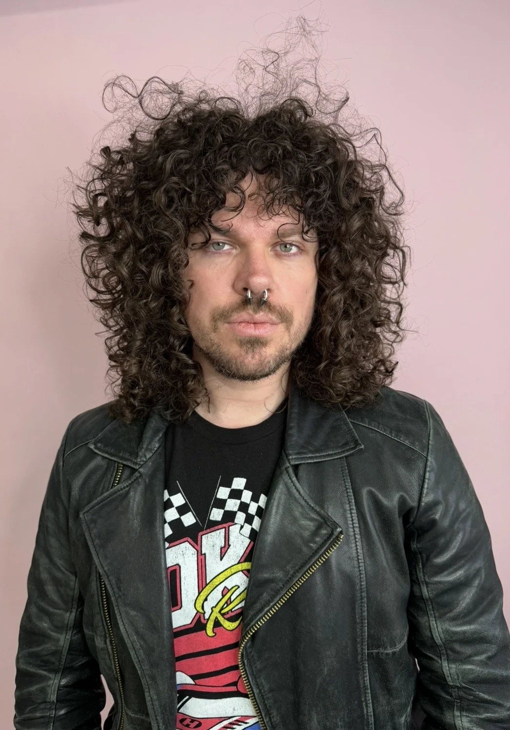 Portrait of a man with long, curly brown hair, light skin, and a septum piercing, wearing a black leather jacket and a graphic t-shirt, standing against a pink background.