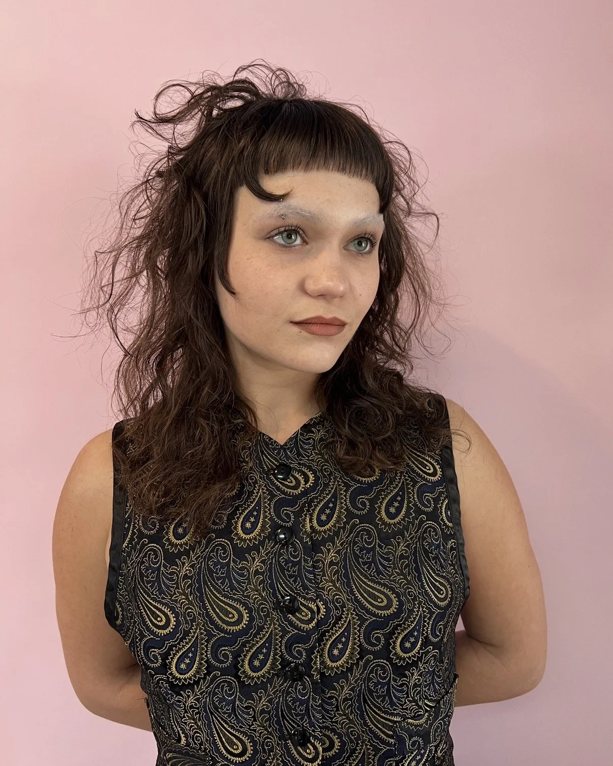A woman with curly brown hair, wearing a black sleeveless top with gold paisley patterns, stands against a pink background.