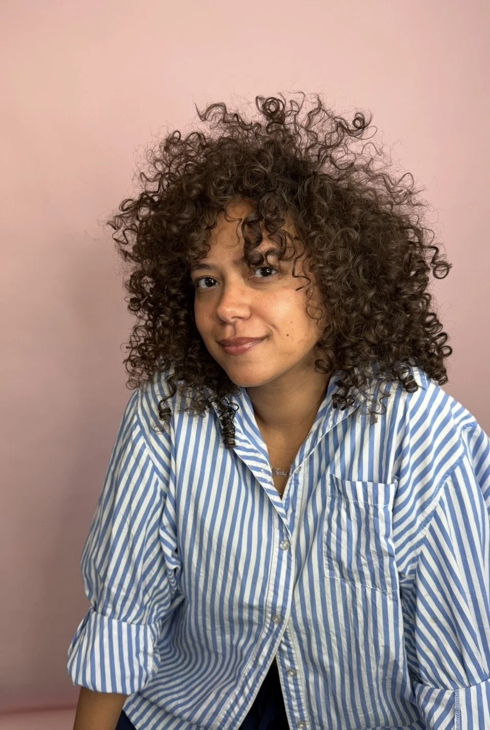 A woman with curly brown hair looking at the camera, wearing a blue and white striped button-up shirt, against a pink background.