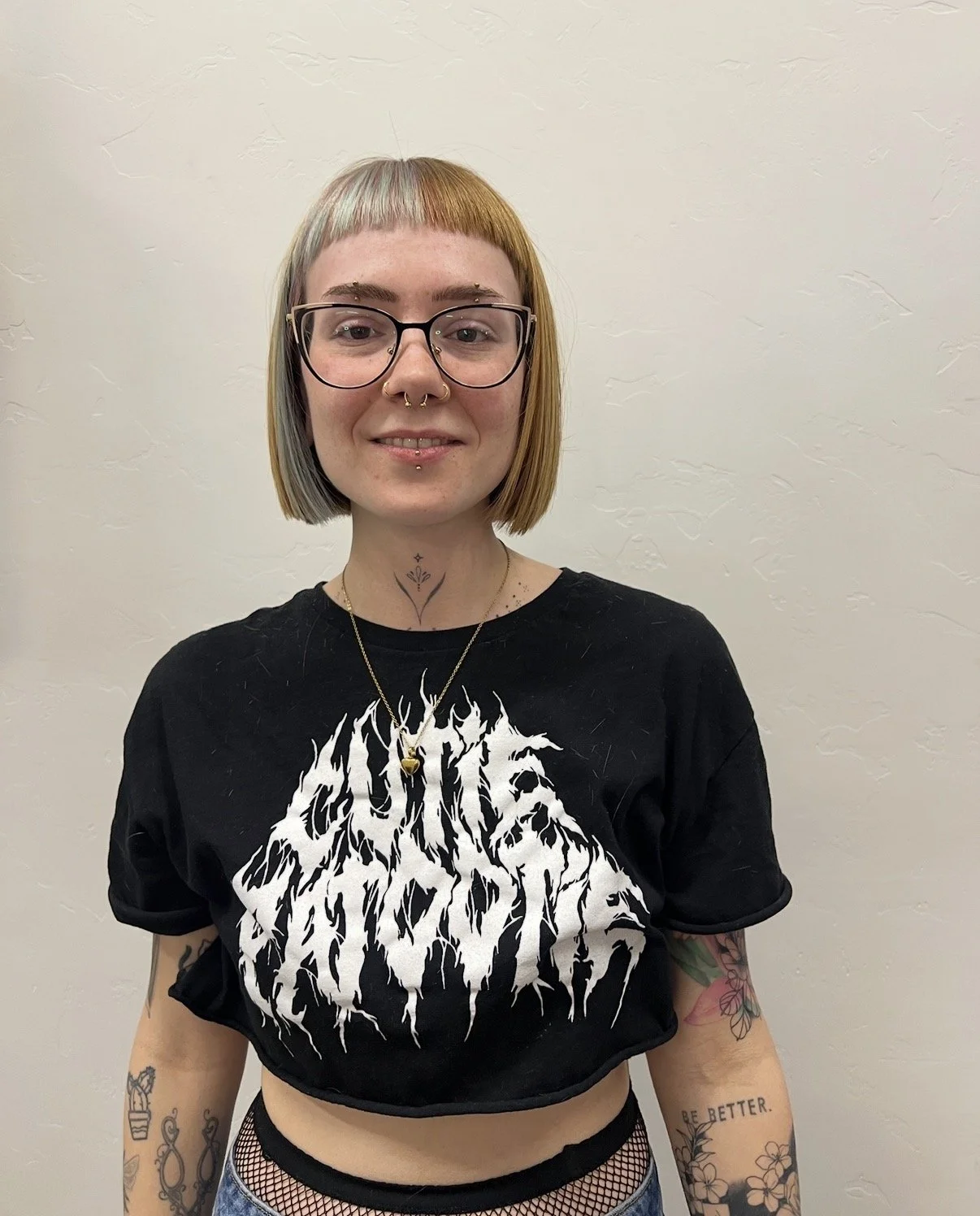 Young woman with short hair, wearing glasses, a black band T-shirt with white graphic design, and tattoos on her arms. She is smiling and standing against a plain wall.