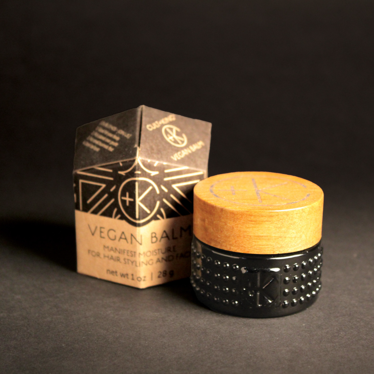Image of vegan balm in a dark glass jar with a wood lid, and the box it comes in.
