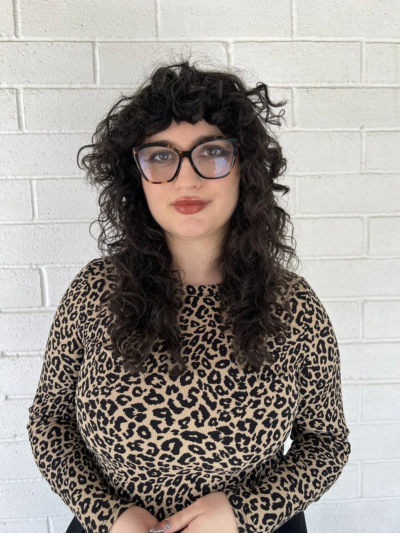 A woman with curly dark hair, glasses, and wearing a leopard print top standing in front of a white brick wall.