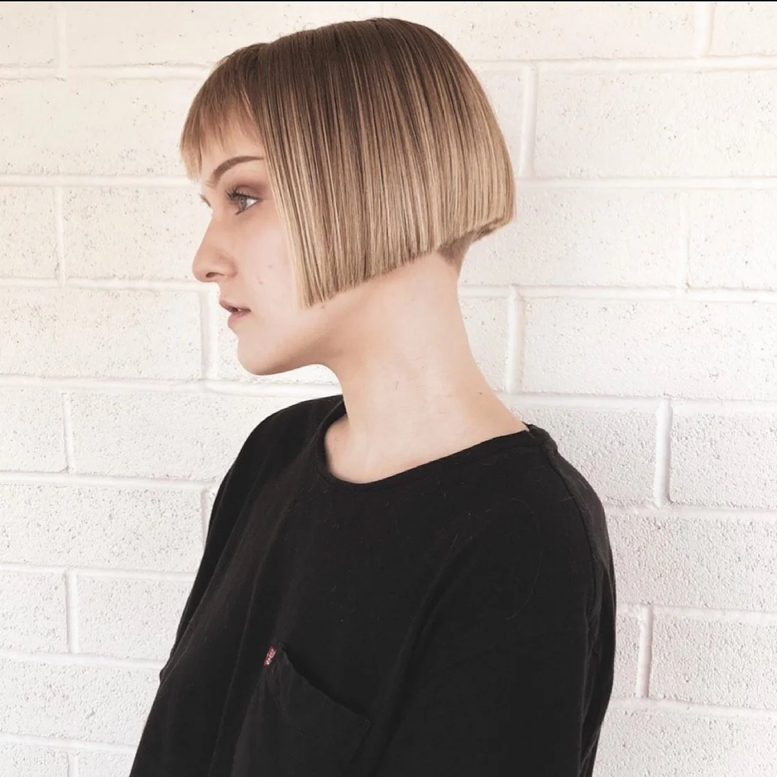 Side profile of a woman with a short bob haircut, light brown hair, and a black shirt standing against a white brick wall.