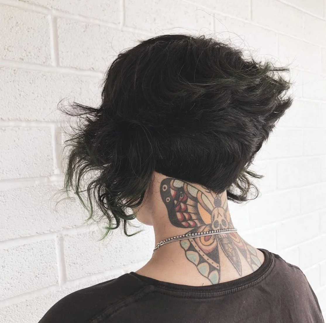 Back of a person with short black and green hair, tattoos on the neck, wearing a chain necklace, against a white brick wall.