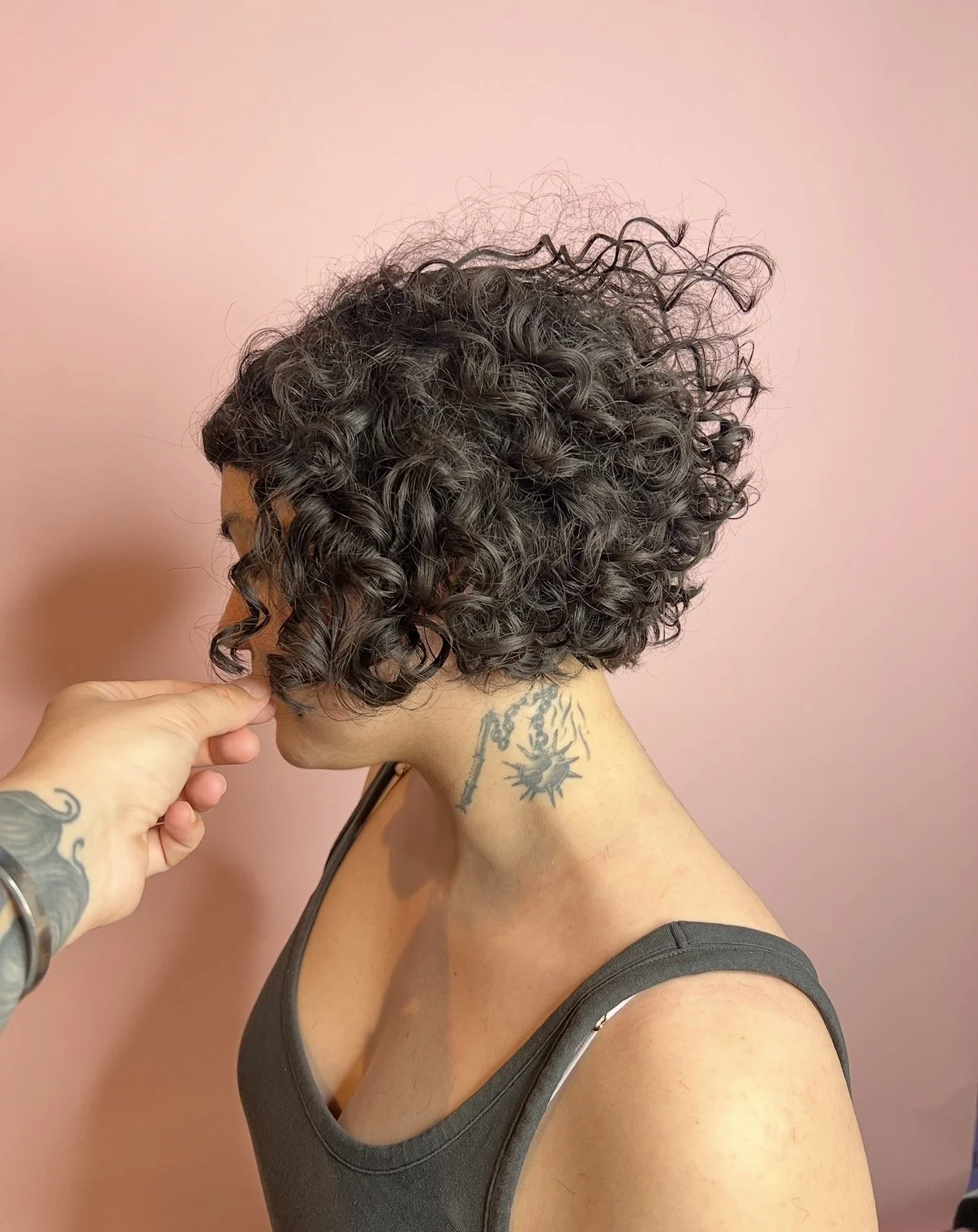 Profile of woman with curly short hairstyle, dark hair, and tattoo on neck, against pink background.