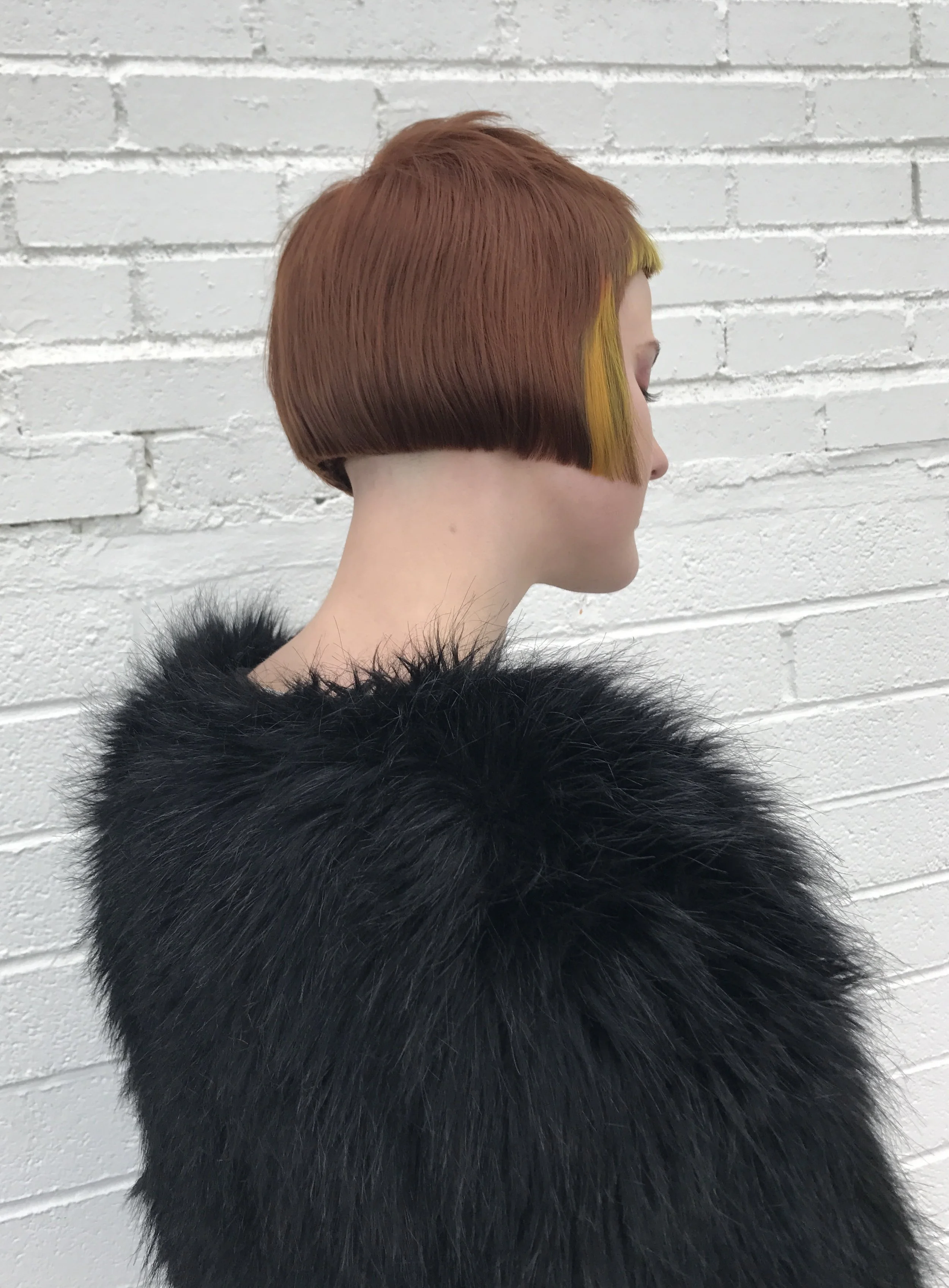 Side view of a woman with a short, geometric hairstyle, featuring brown and yellow hair colors, wearing a black fur coat, against a white brick wall.