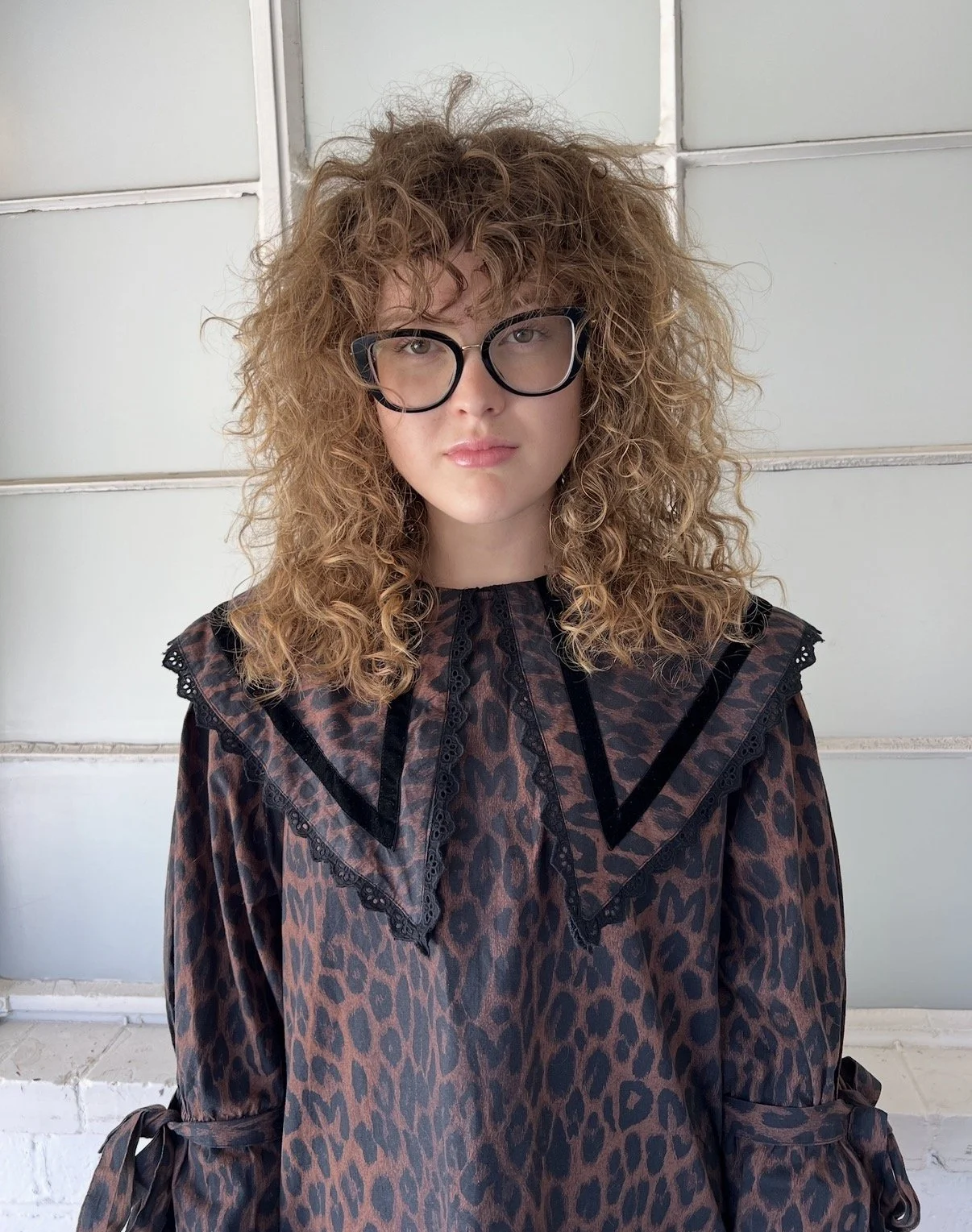 Young woman with curly red hair and glasses wearing a leopard print blouse with black lace accents, standing in front of a light-colored wall.