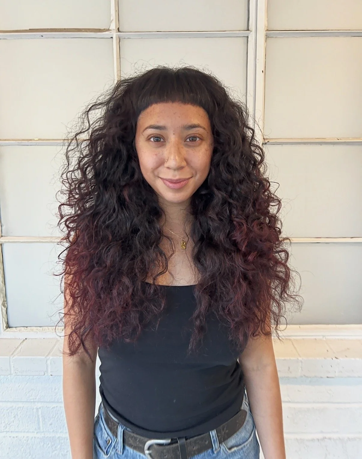 A young woman with long curly dark hair and freckles, smiling at the camera, wearing a black t-shirt and blue jeans, standing in front of a white brick wall.