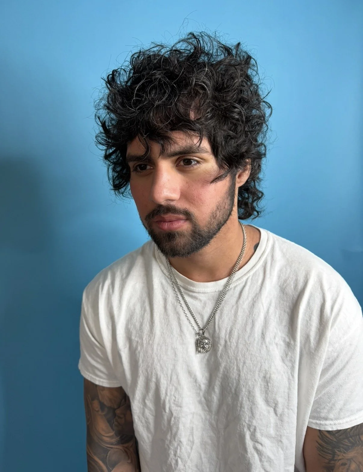 A young man with messy curly black hair, a beard, and tattoos on his arms, wearing a white t-shirt and silver necklaces, poses against a blue background.
