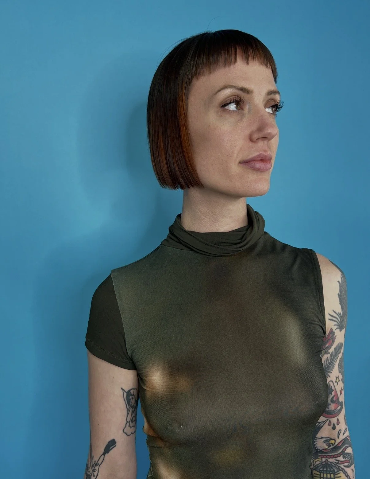 Portrait of a woman with short brown hair and tattoos wearing a sheer black turtleneck shirt against a blue background.