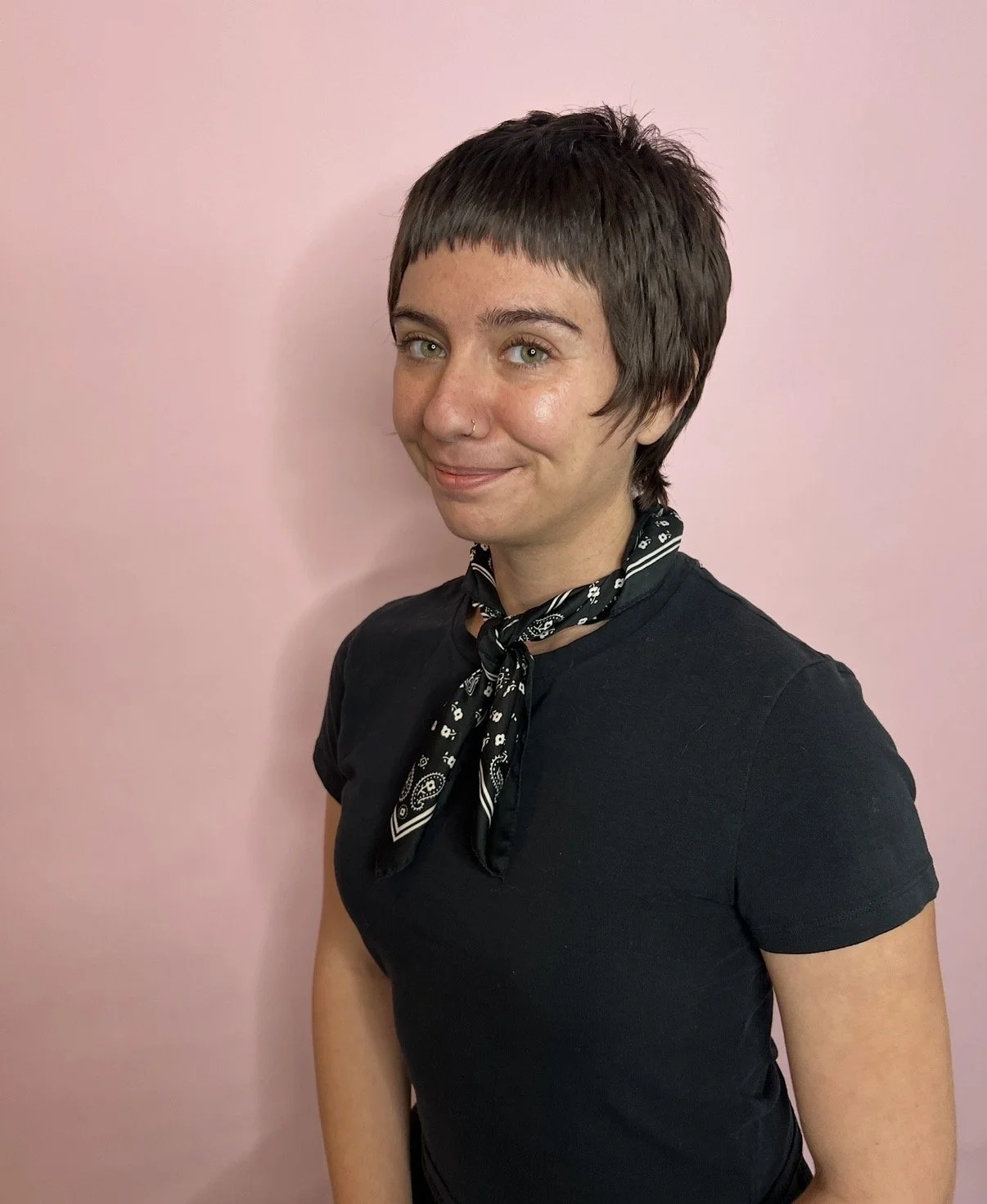 A young person with short dark hair, light skin, and green eyes, wearing a black t-shirt and a black bandana with white patterns around their neck, standing against a pink background.