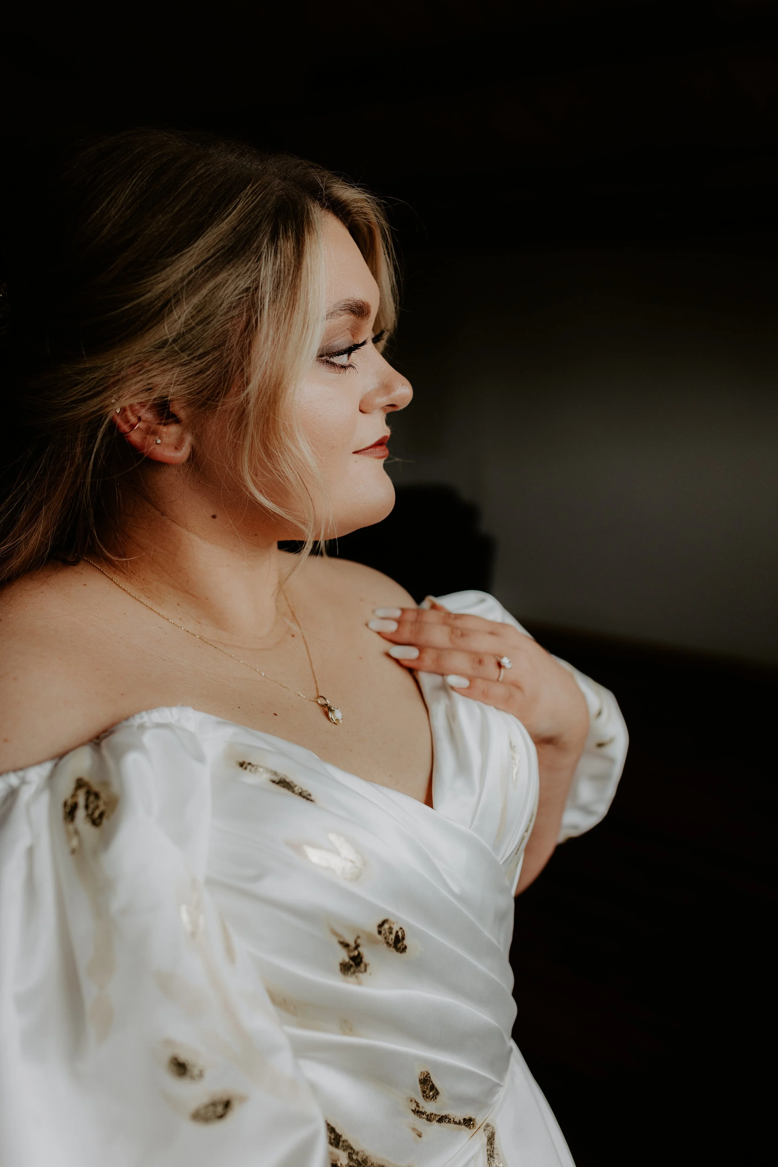 A woman in a white off-shoulder dress looking to the side with her hand on her chest, wearing jewelry and makeup, against a dark background.