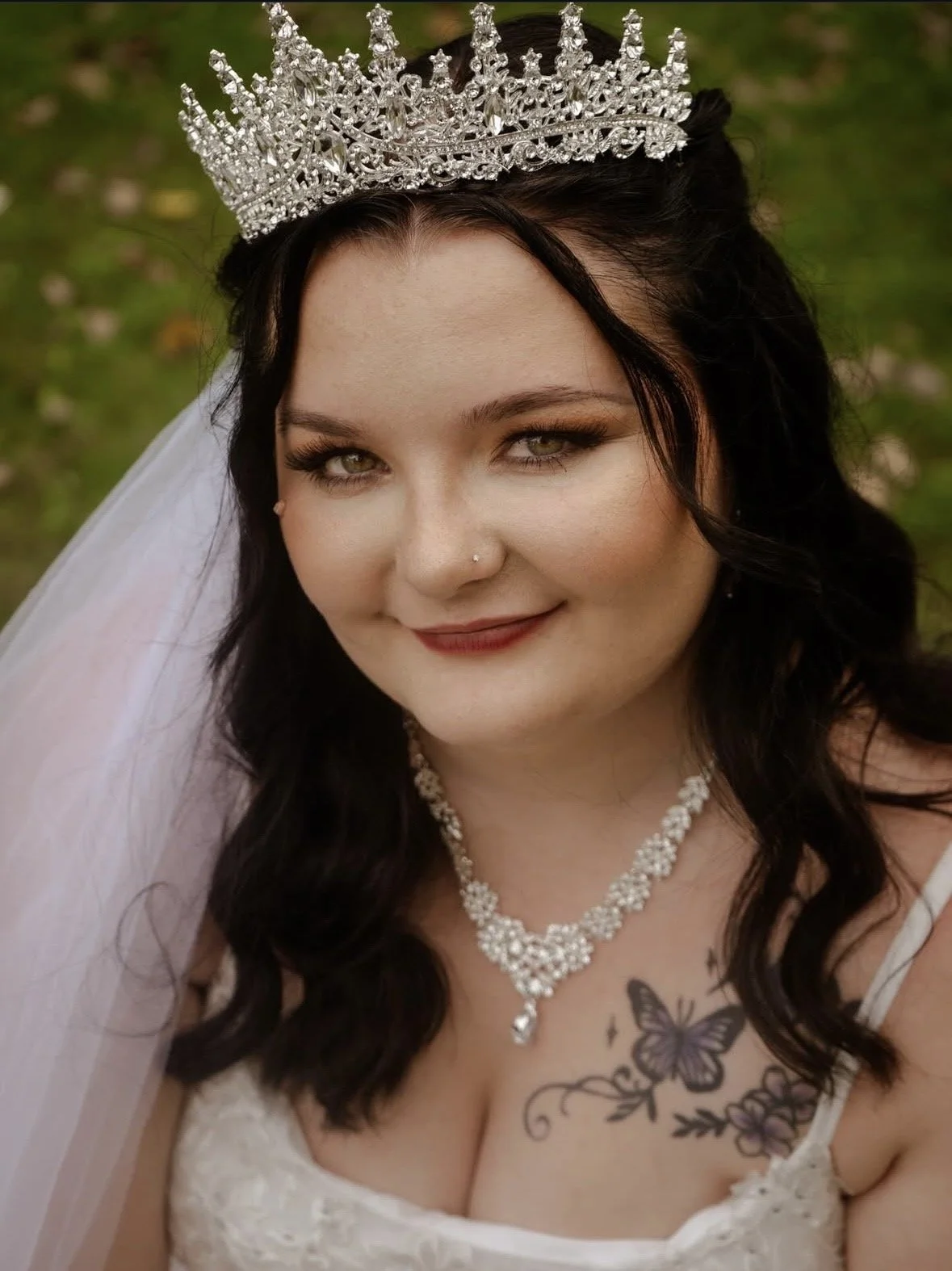 A woman with dark hair, wearing a tiara, white dress, and pearl necklace, smiling outdoors, with a butterfly tattoo on her chest.