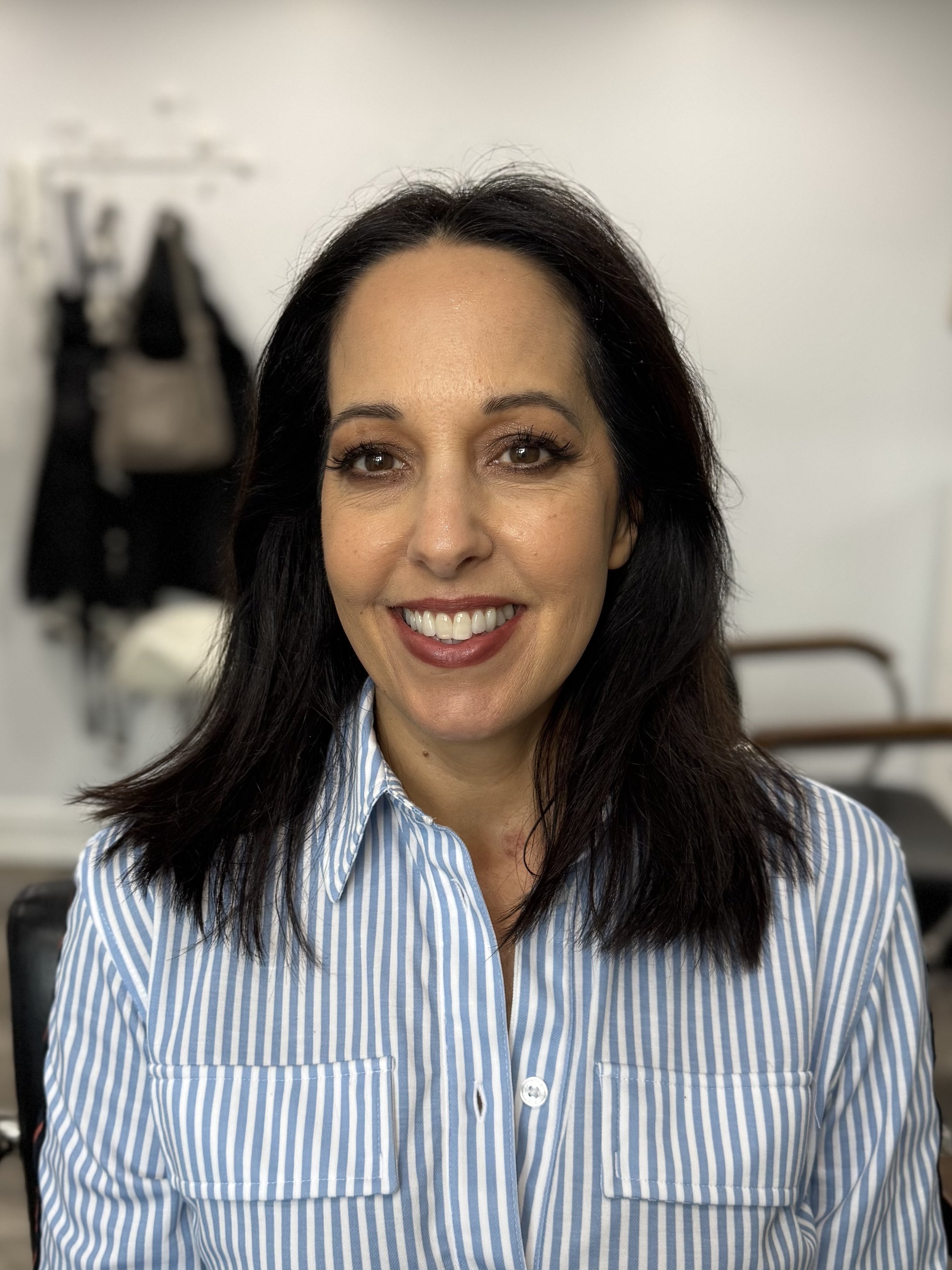 woman smiling with dark hair and brown eyes, wearing a blue and white striped button-up shirt, in a room with blurred background including bags hanging on a wall and furniture