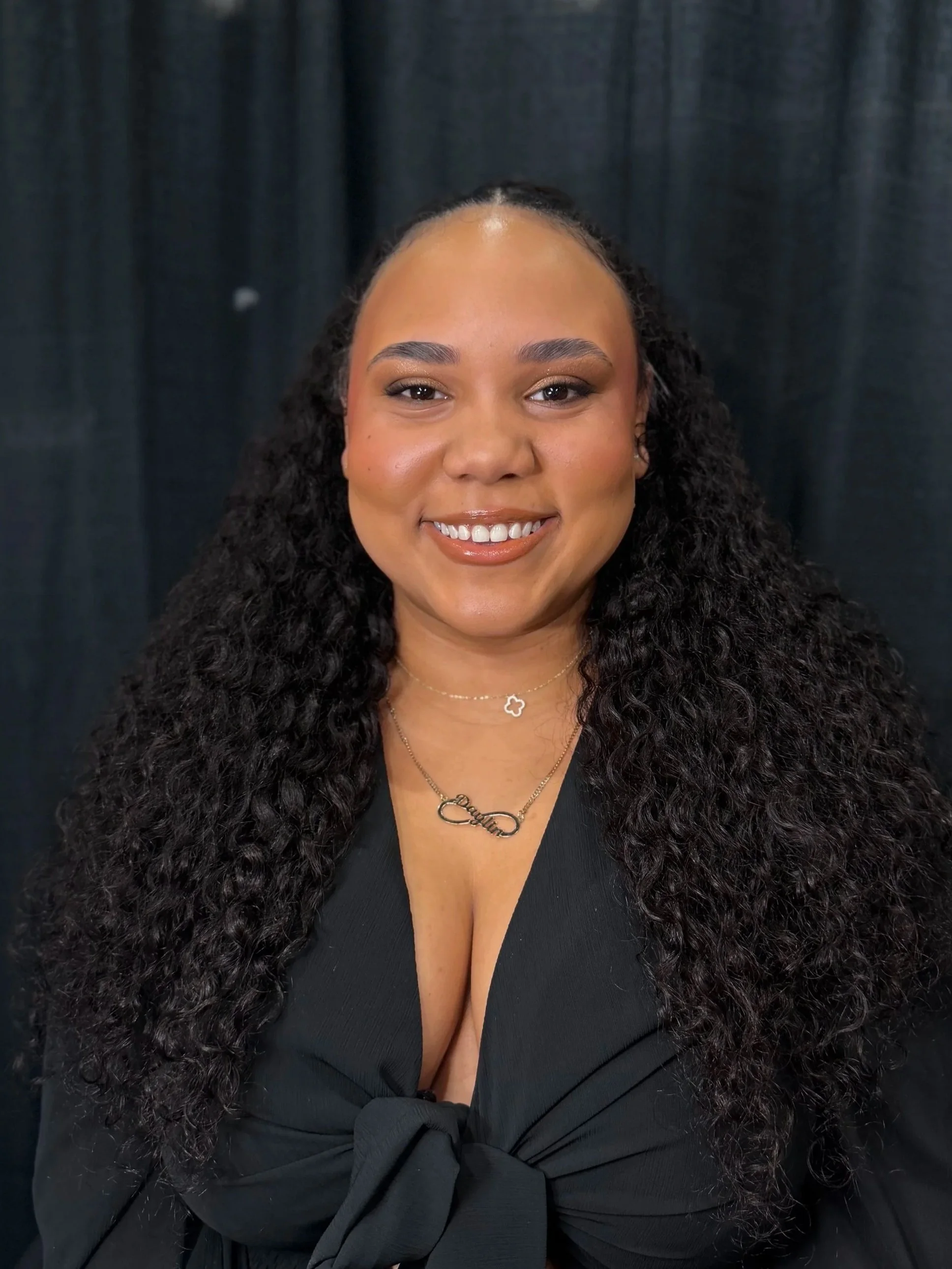 A smiling woman with long, curly black hair, wearing a black top with a knot at the front, gold necklaces, and a black background.