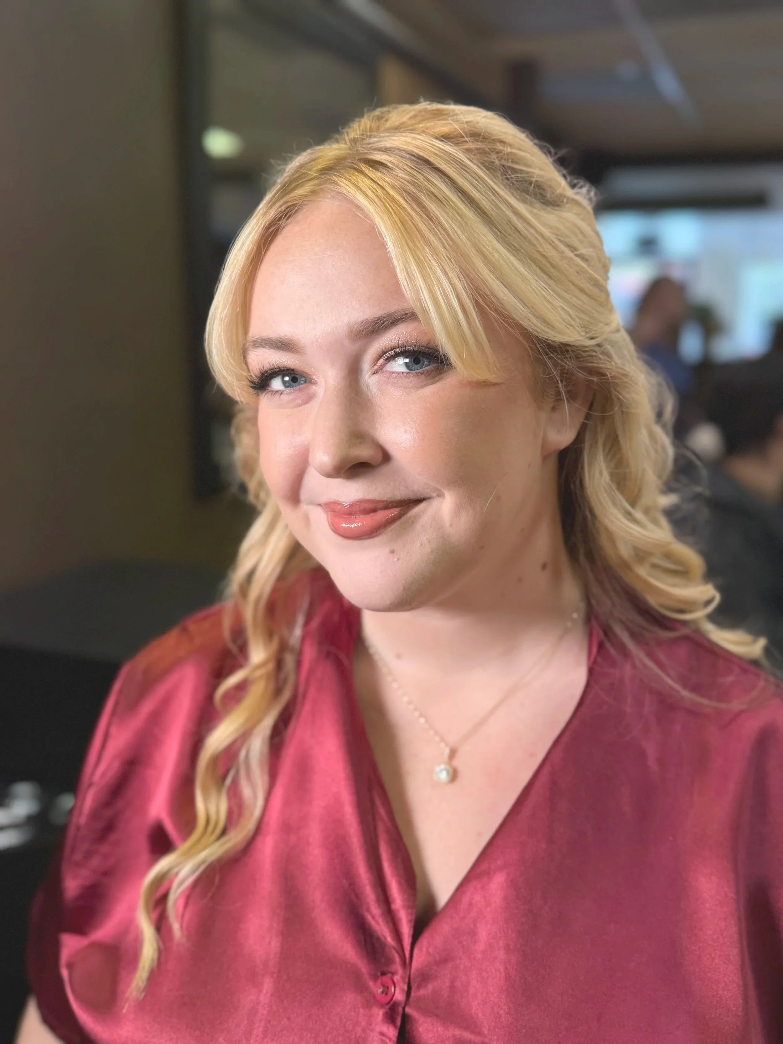 A smiling woman with blonde wavy hair, blue eyes, and wearing a maroon satin blouse and a delicate necklace, standing indoors with a blurred background.