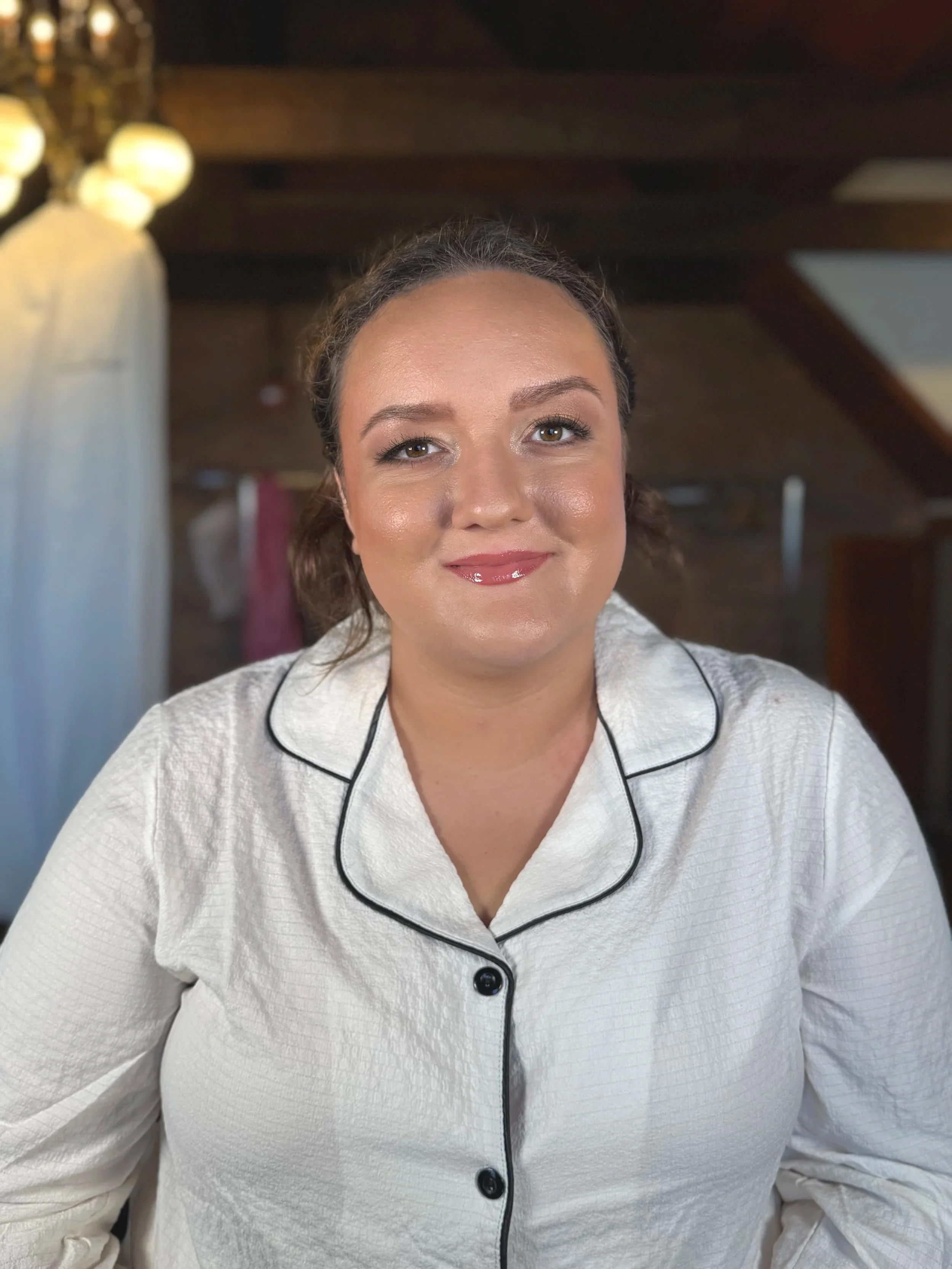 A woman with medium-length brown hair, light makeup, wearing a white textured pajama top with black piping, smiling at the camera, in a cozy, wooden interior with warm lighting.