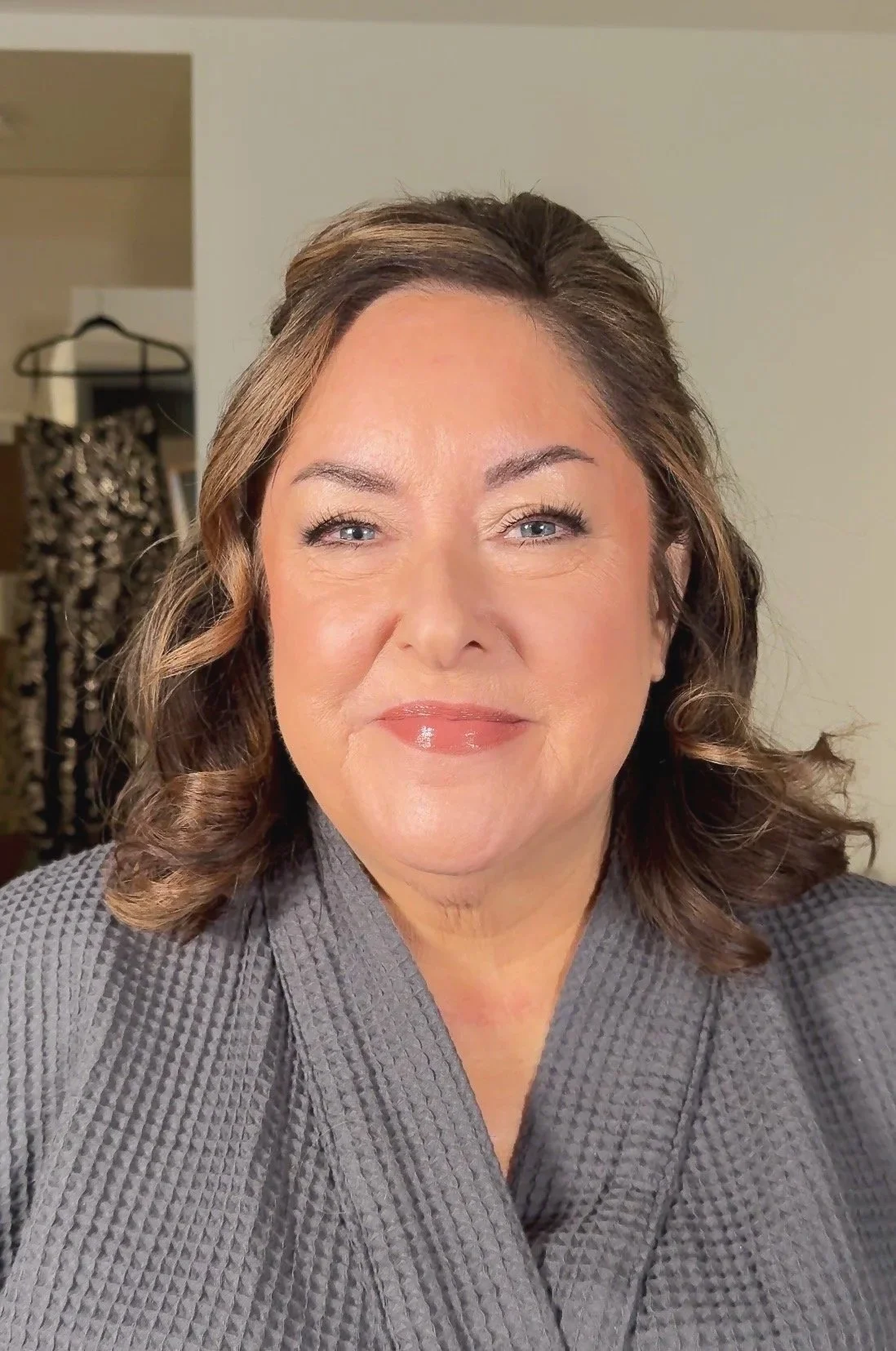 A woman with shoulder-length brown hair, wearing a gray textured kimono-style top, smiling at the camera. The background shows an open closet with some hangers and clothing.