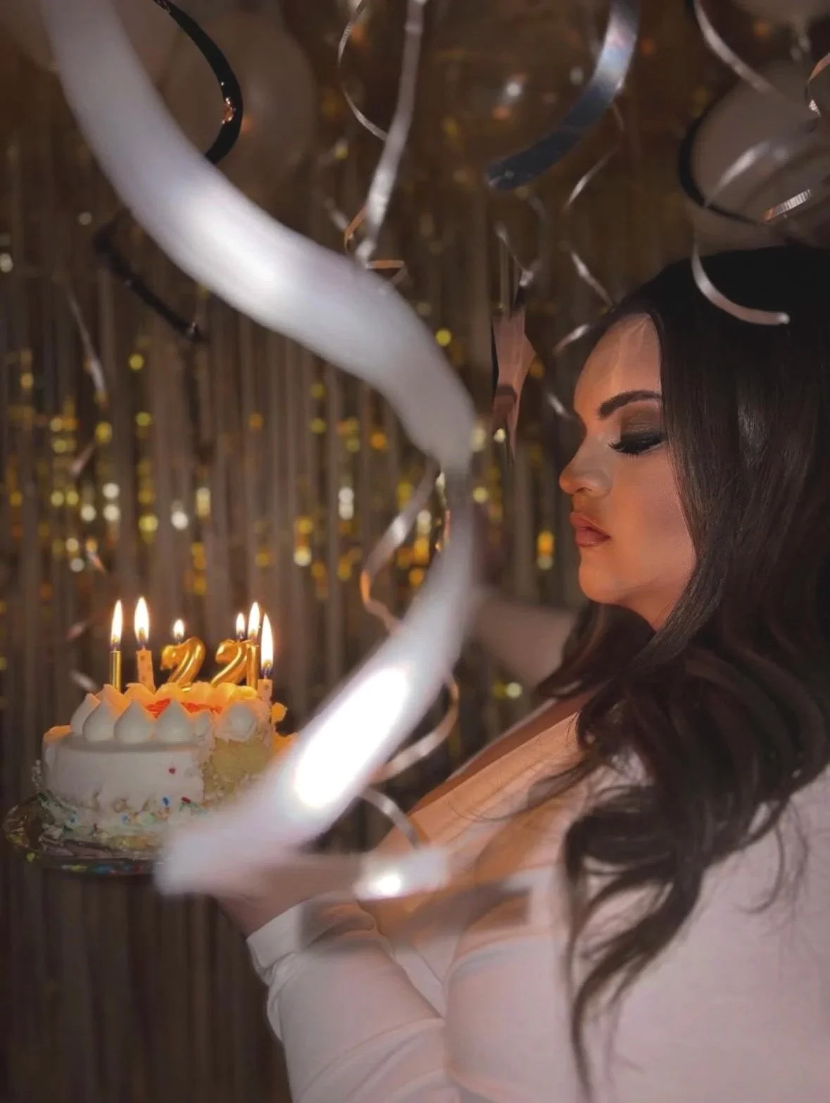 A woman with long dark hair and makeup celebrates her birthday with a cake that has lit candles numbered 1, 2, and 2, surrounded by silver and white streamers and balloons in the background.