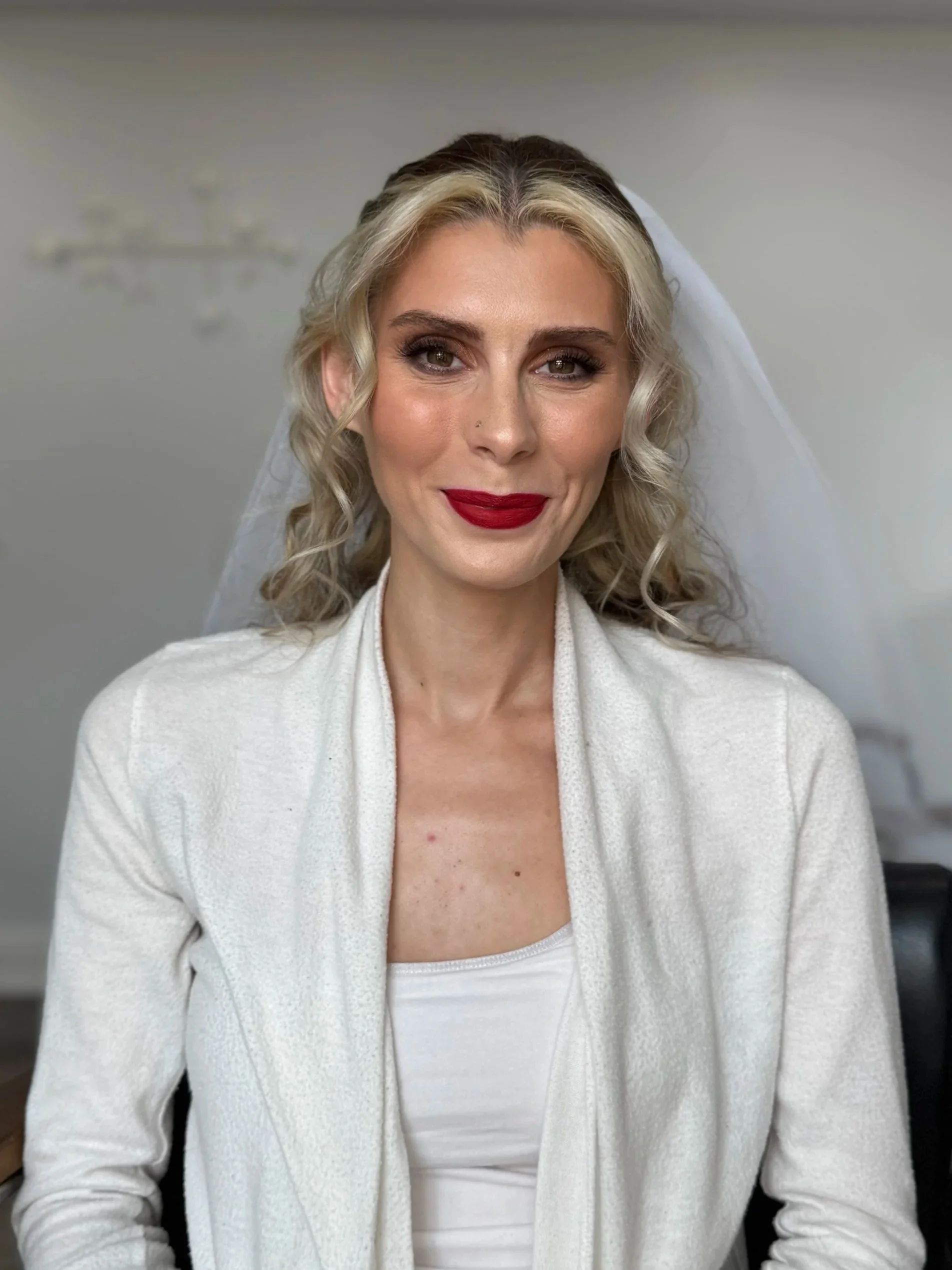 A woman with curly blonde hair wearing a white cardigan, white top, red lipstick, and makeup, smiling in a well-lit room with a blurred background.