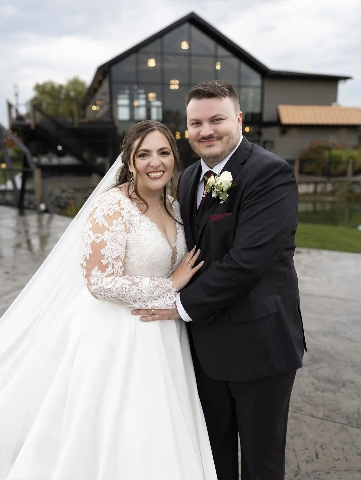 A newlywed couple posing outdoors in front of a modern building with large glass windows. The bride is wearing a white lace wedding dress and veil, and the groom is in a black suit with a white shirt and boutonniere. They are smiling at the camera.