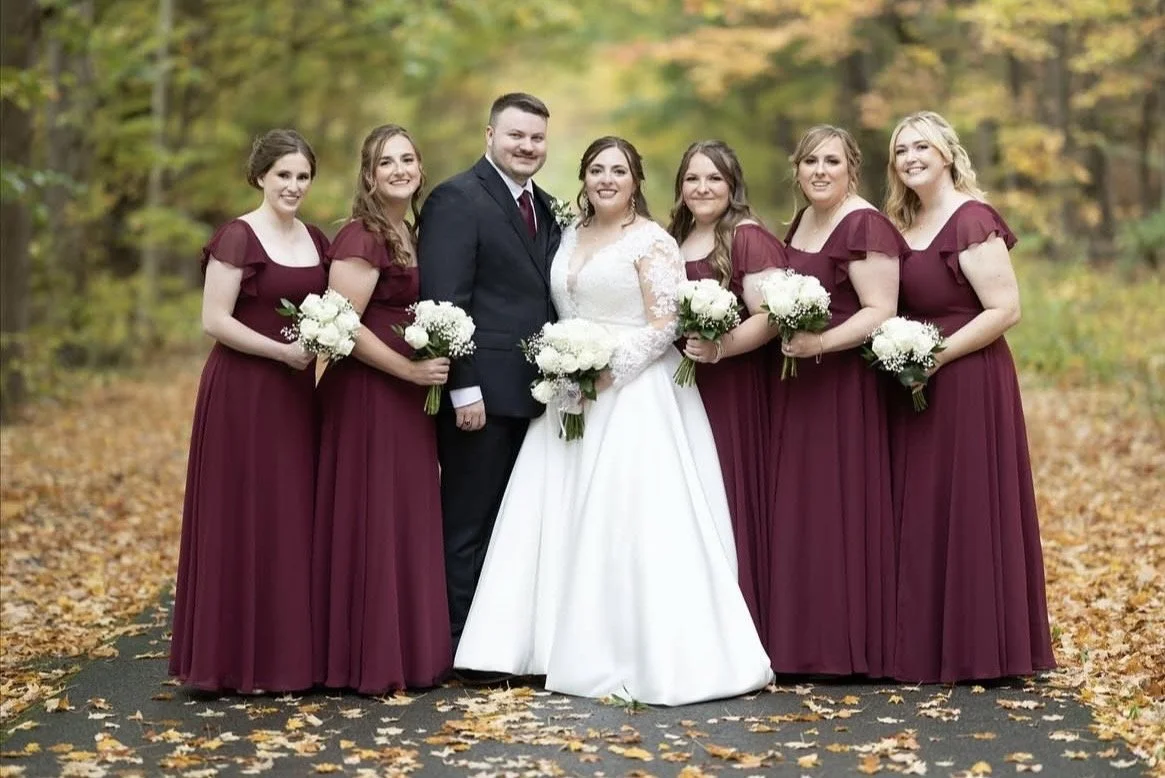A wedding party with seven people, including a bride in a white gown, a groom in a dark suit, and five bridesmaids in matching burgundy dresses, standing outdoors on a leaf-covered path in a forest with autumn foliage.