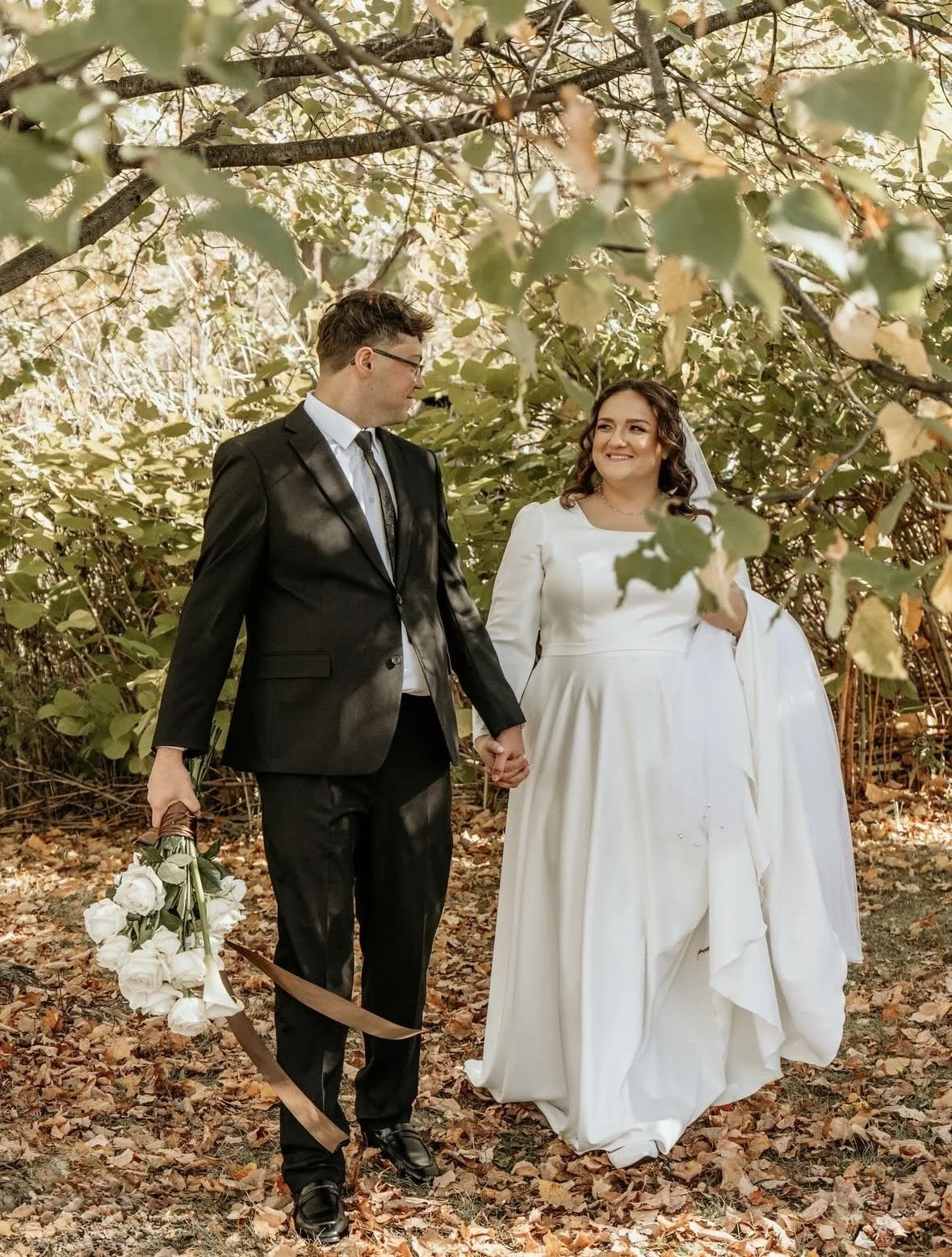 A bride and groom walking hand in hand through a wooded area, holding a bouquet of white roses, on their wedding day.