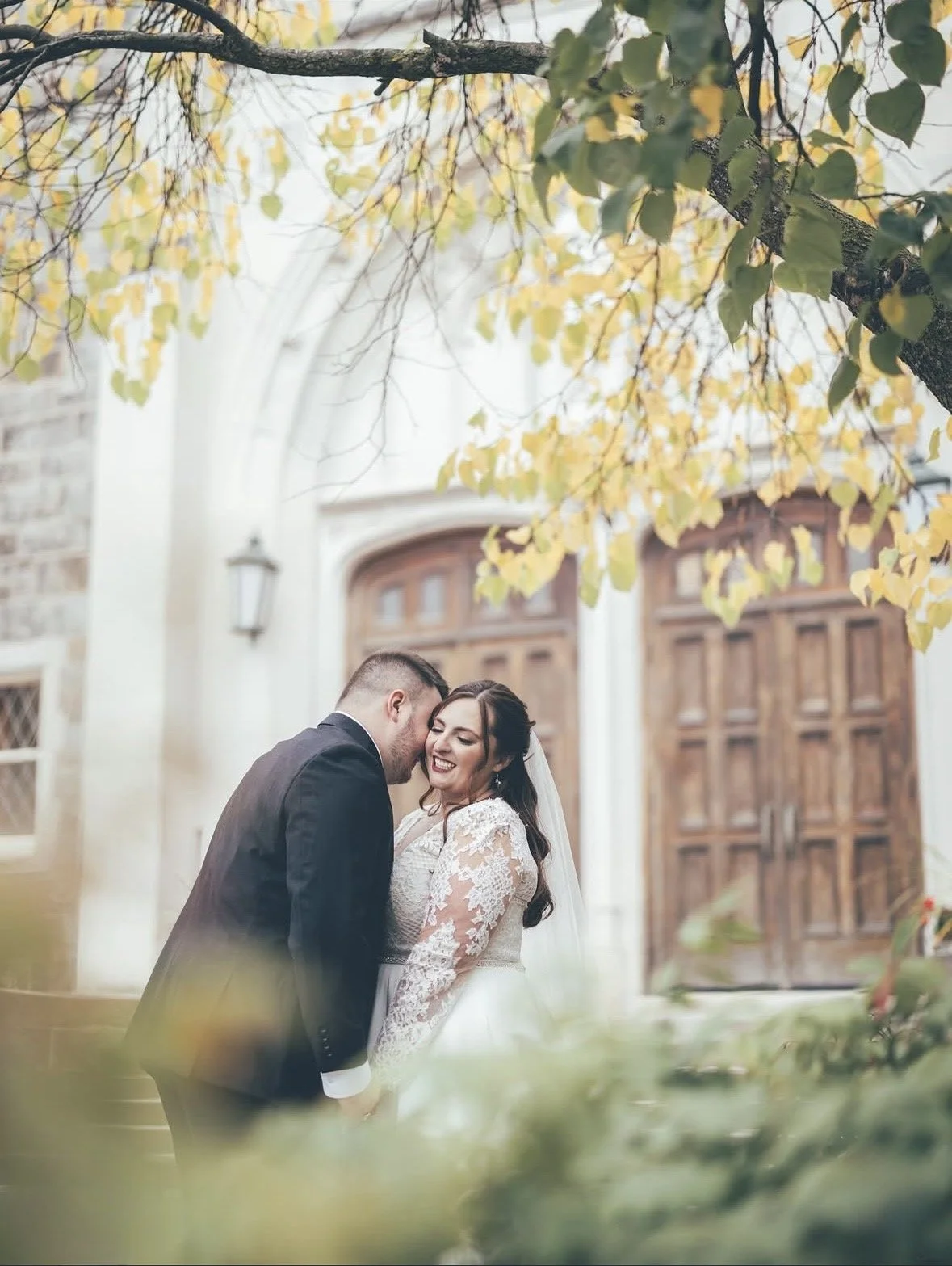 A happy bride and groom sharing an intimate moment outdoors during a wedding, with a church building and large wooden doors in the background and yellow autumn leaves overhead.