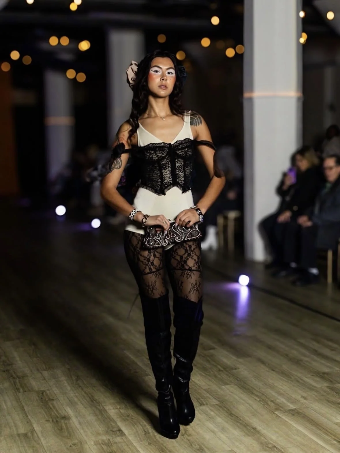 Woman walking on runway in a fashion show, wearing black lace pants, a beige top with black lace overlay, and holding a small clutch purse, with audience seated in the background.