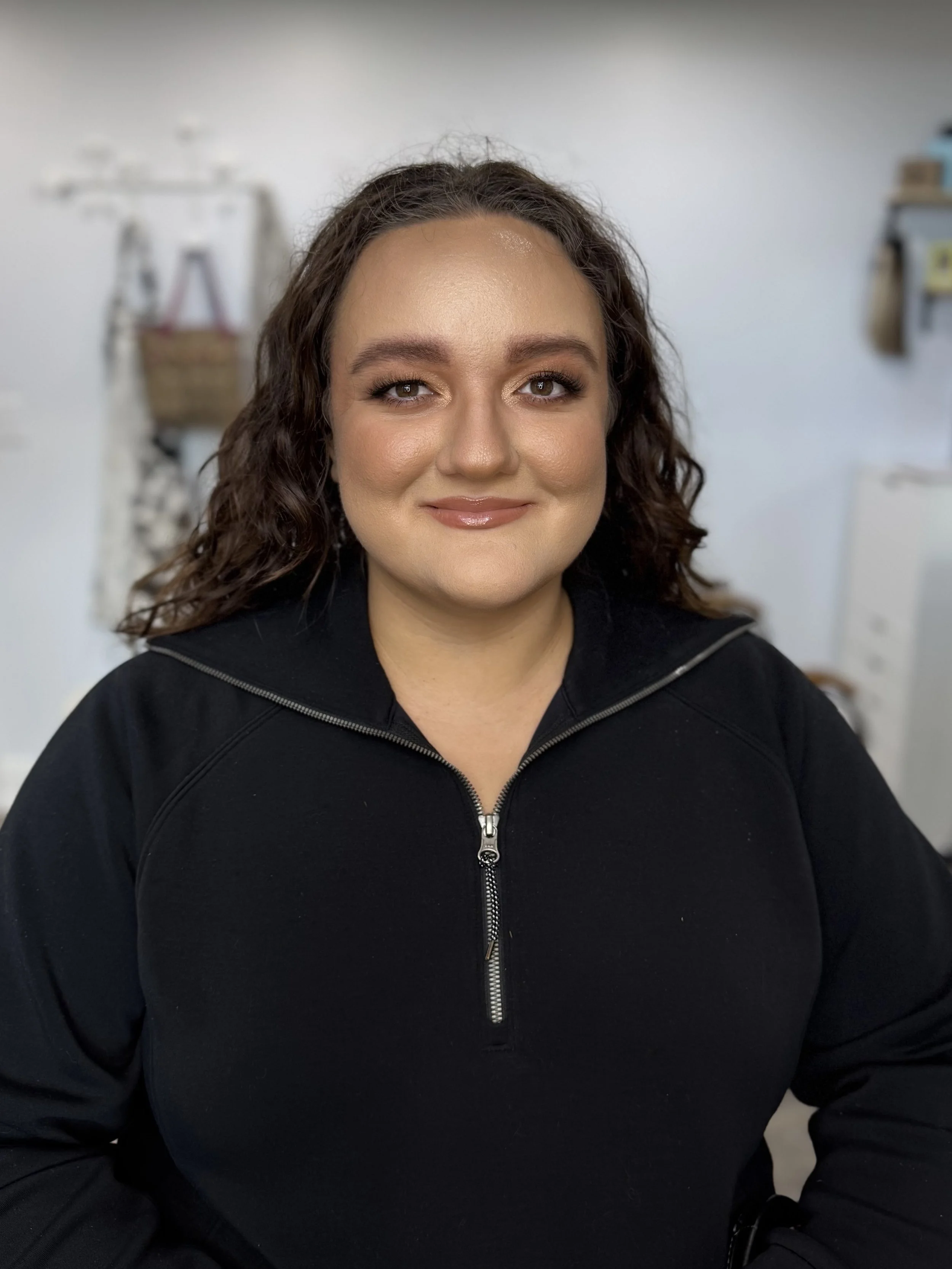A woman with curly brown hair and makeup, wearing a black zip-up jacket, smiling at the camera in an indoor setting.