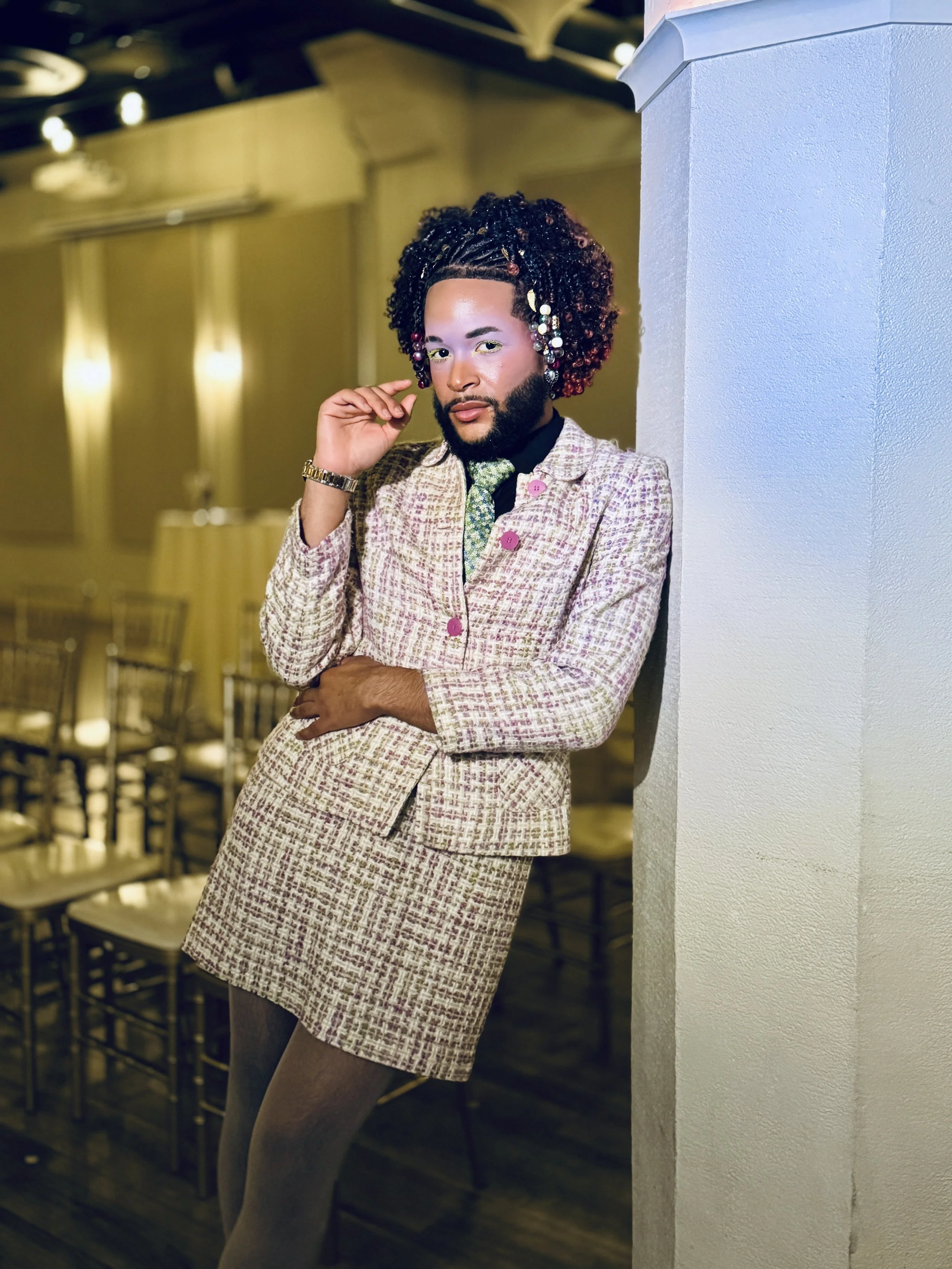 A person with curly hair styled with beads and accessories, wearing a patterned blazer and skirt, standing against a wall in an indoor setting with chairs in the background.
