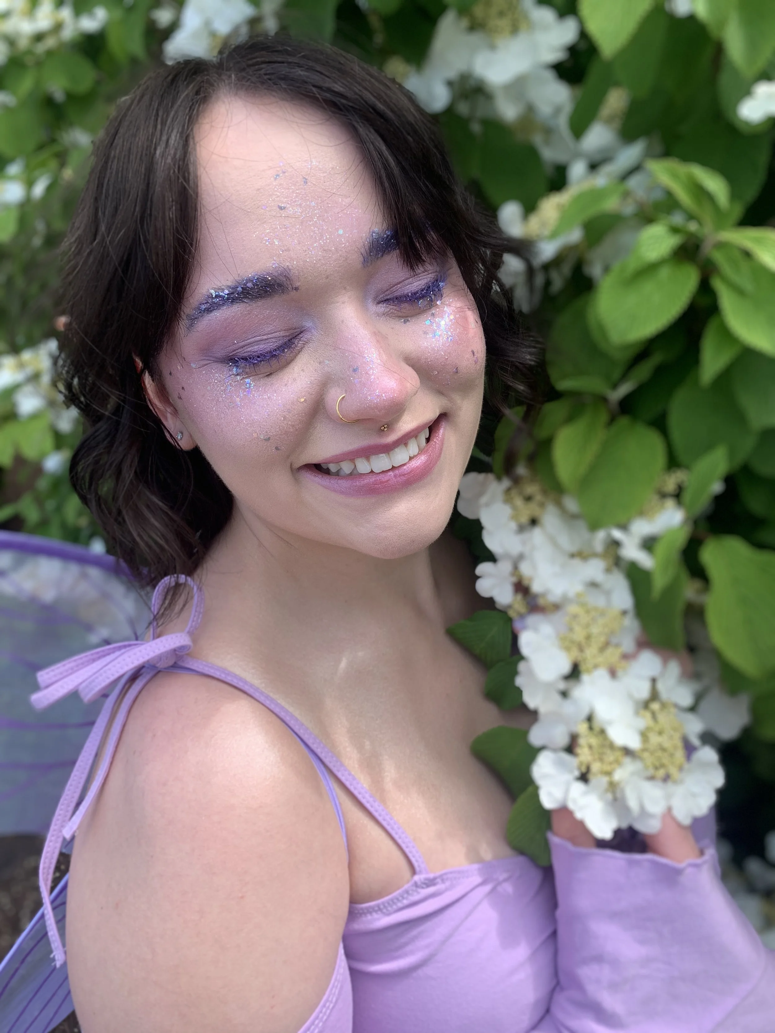 A woman with short dark hair and fairy wings, wearing purple makeup and glitter, holding a bunch of white flowers with green leaves, smiling with closed eyes outdoors.